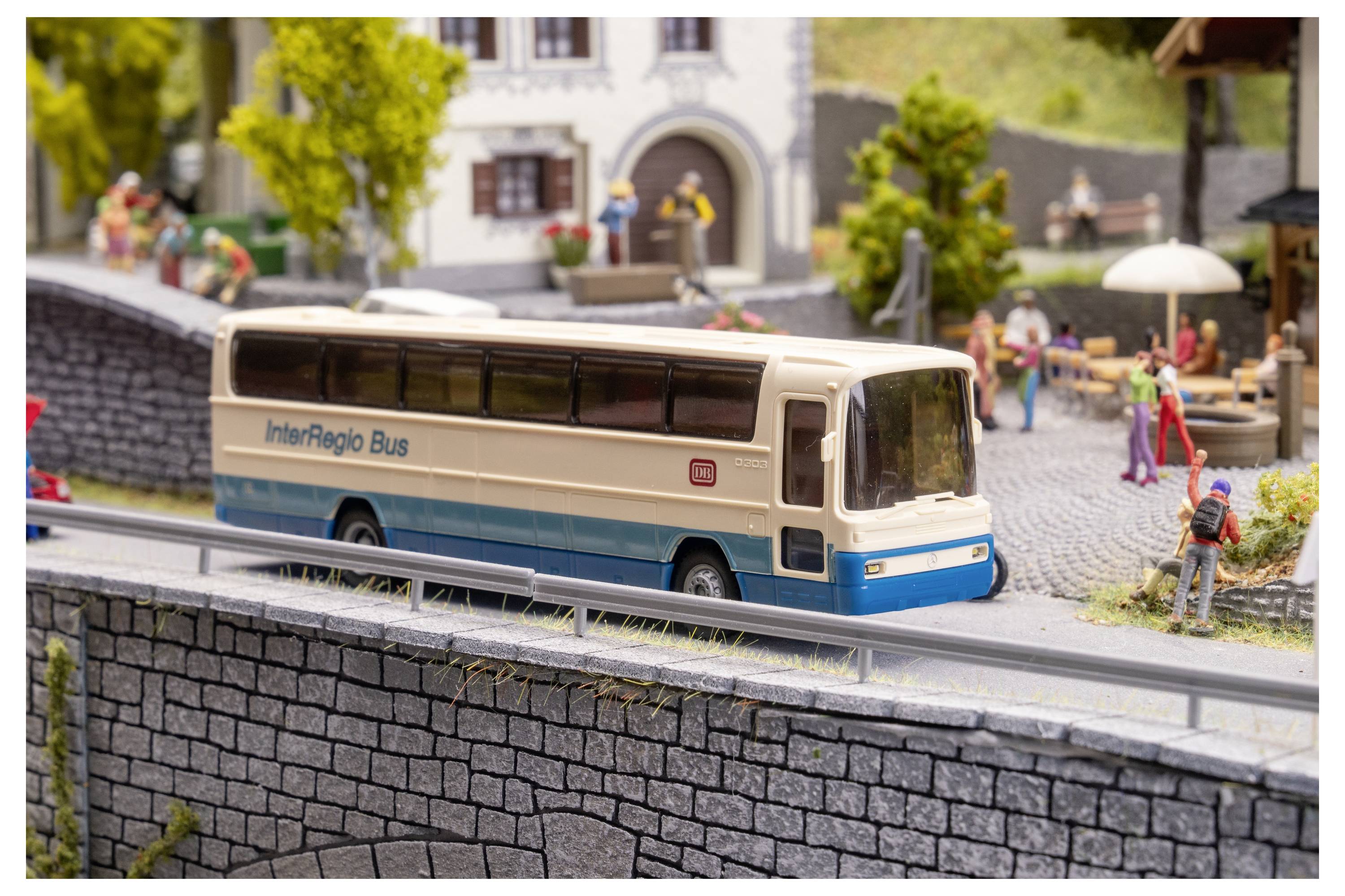 Modellbus im Fernverkehr auf einer Straße in einem Miniatur-Dorf, vorbei an Fußgängern und Häusern in einem detaillierten Diorama.