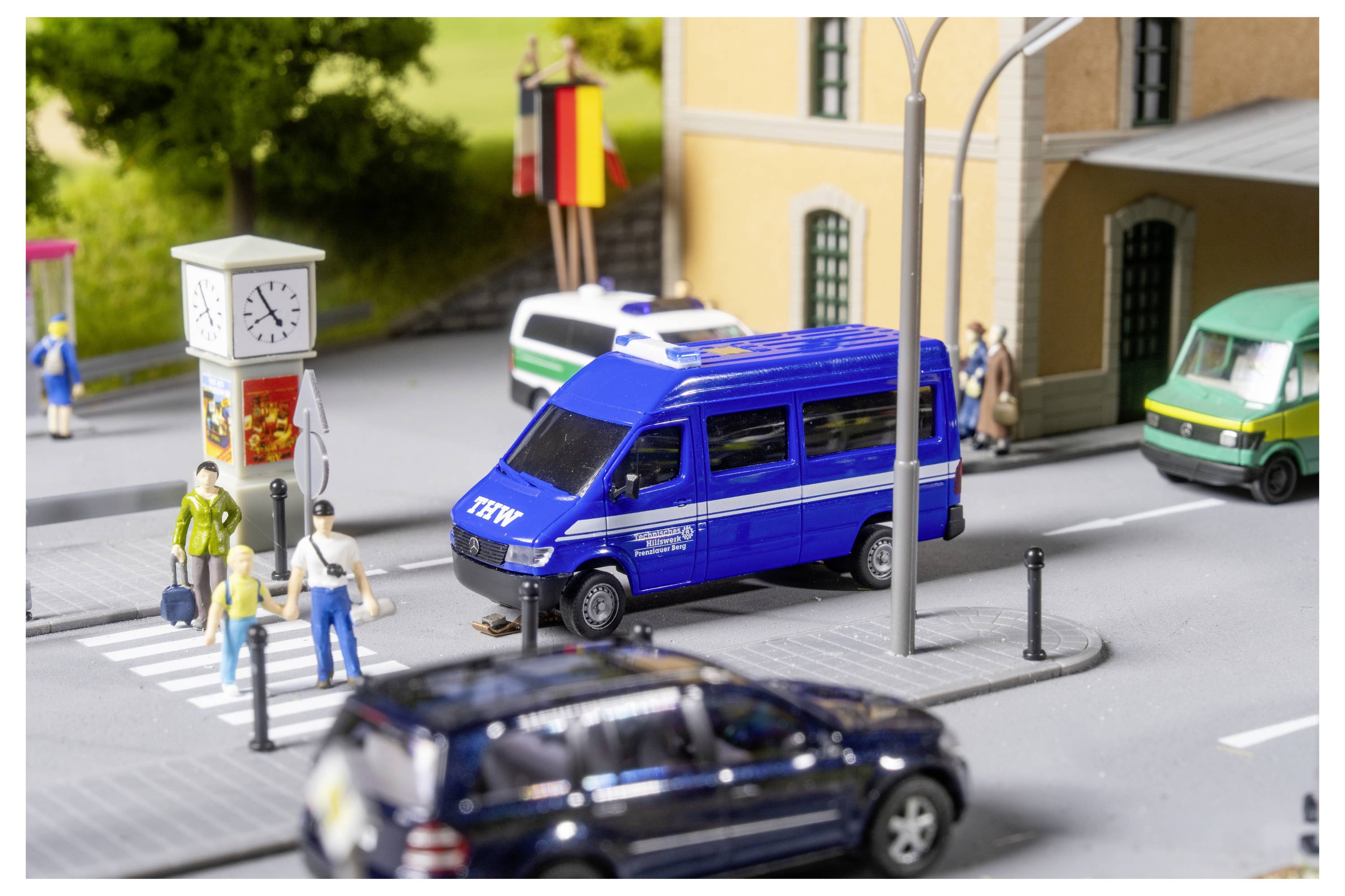 Miniaturstraßenszene mit einem blauen Polizeiwagen, der an einem Zebrastreifen neben Fußgängern hält, ein Uhr-Kiosk und Gebäude; ein unscharfes Auto fährt im Vordergrund vorbei.