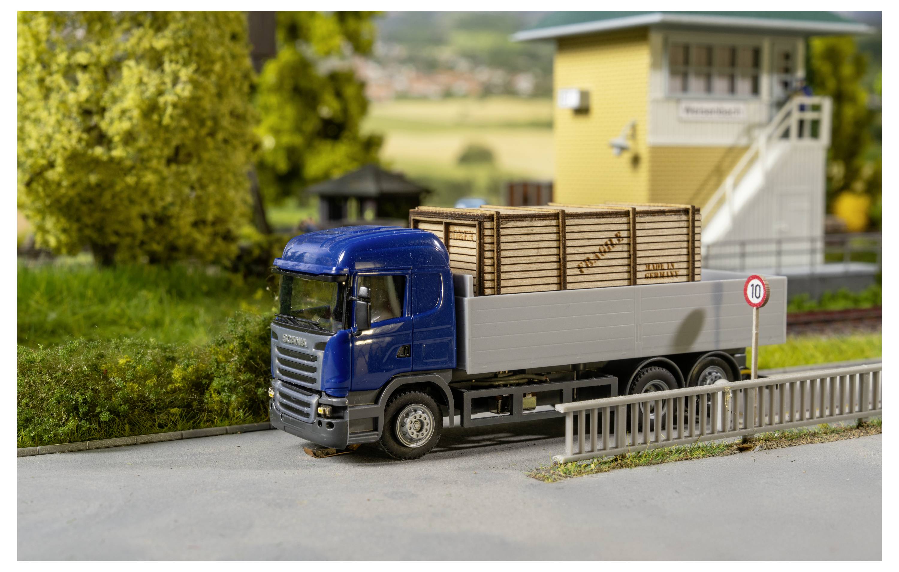 Blauer Pritschenwagen mit Holzkisten auf einer Straße neben einem Geschwindigkeitsbegrenzungsschild mit der Zahl 10, im Hintergrund ein gelbes Gebäude und Grünflächen.