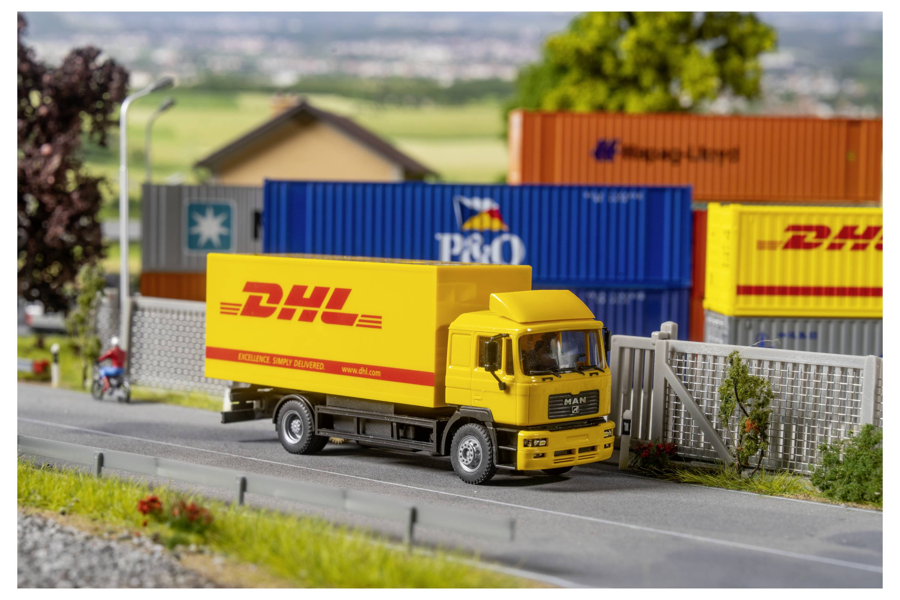 Gelber DHL-Lieferwagen, der auf einer Straße neben gestapelten Versandcontainern in einer Miniaturszene eines Frachtplatzes fährt.