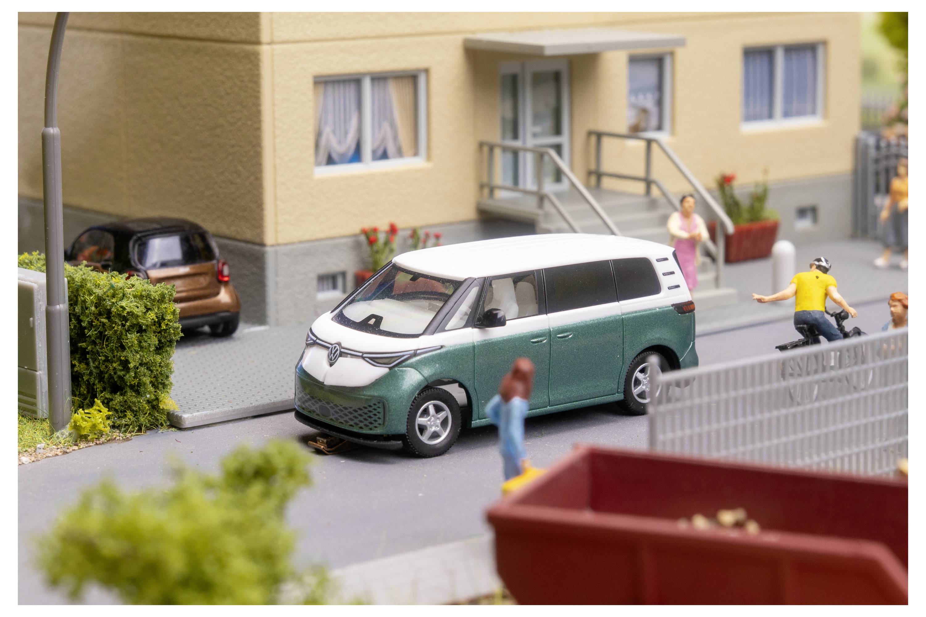 Miniaturstraßenszene mit einem grün-weißen Transporter, der neben einem Gebäude geparkt ist, einem Radfahrer, Fußgängern und einem geparkten Auto im Hintergrund.