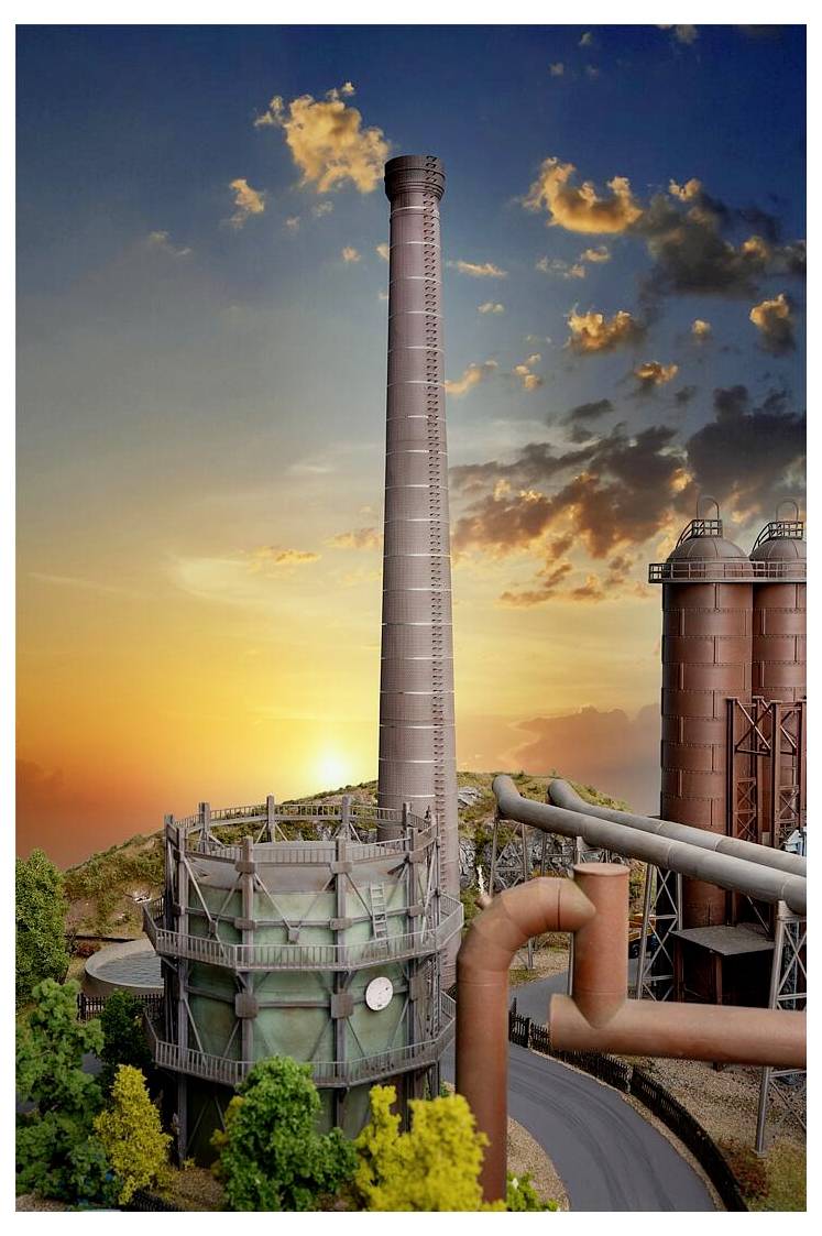 Industrieanlage mit hohem Schornstein, Silos und großen Rohren bei Sonnenuntergang, von oben betrachtet mit Bäumen im Vordergrund.