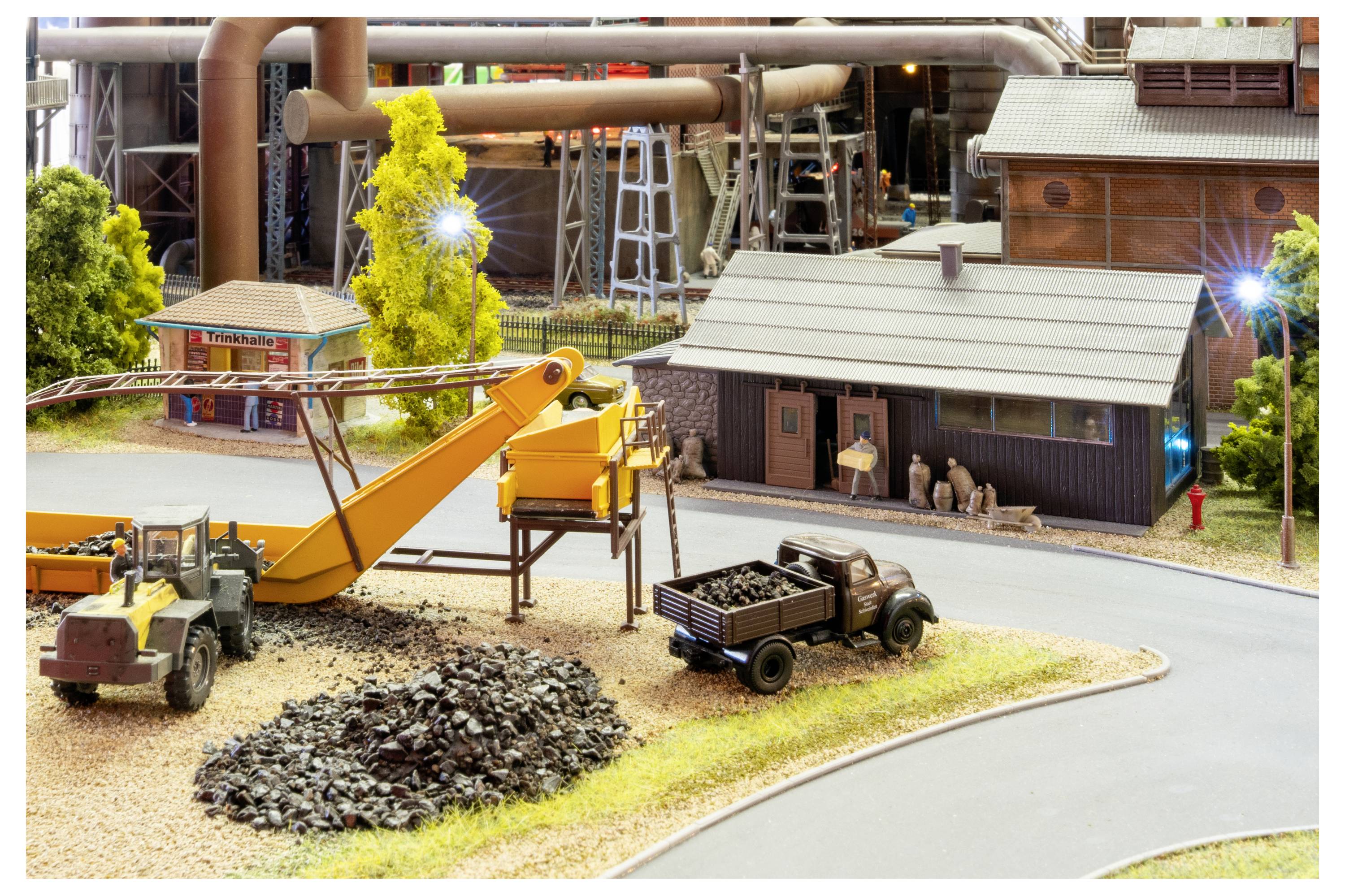 Modellbaustelle mit Bagger, Kipplaster, Kieshaufen und kleinen Gebäuden neben einer Straße auf einer Miniatur-Industrieanlage.