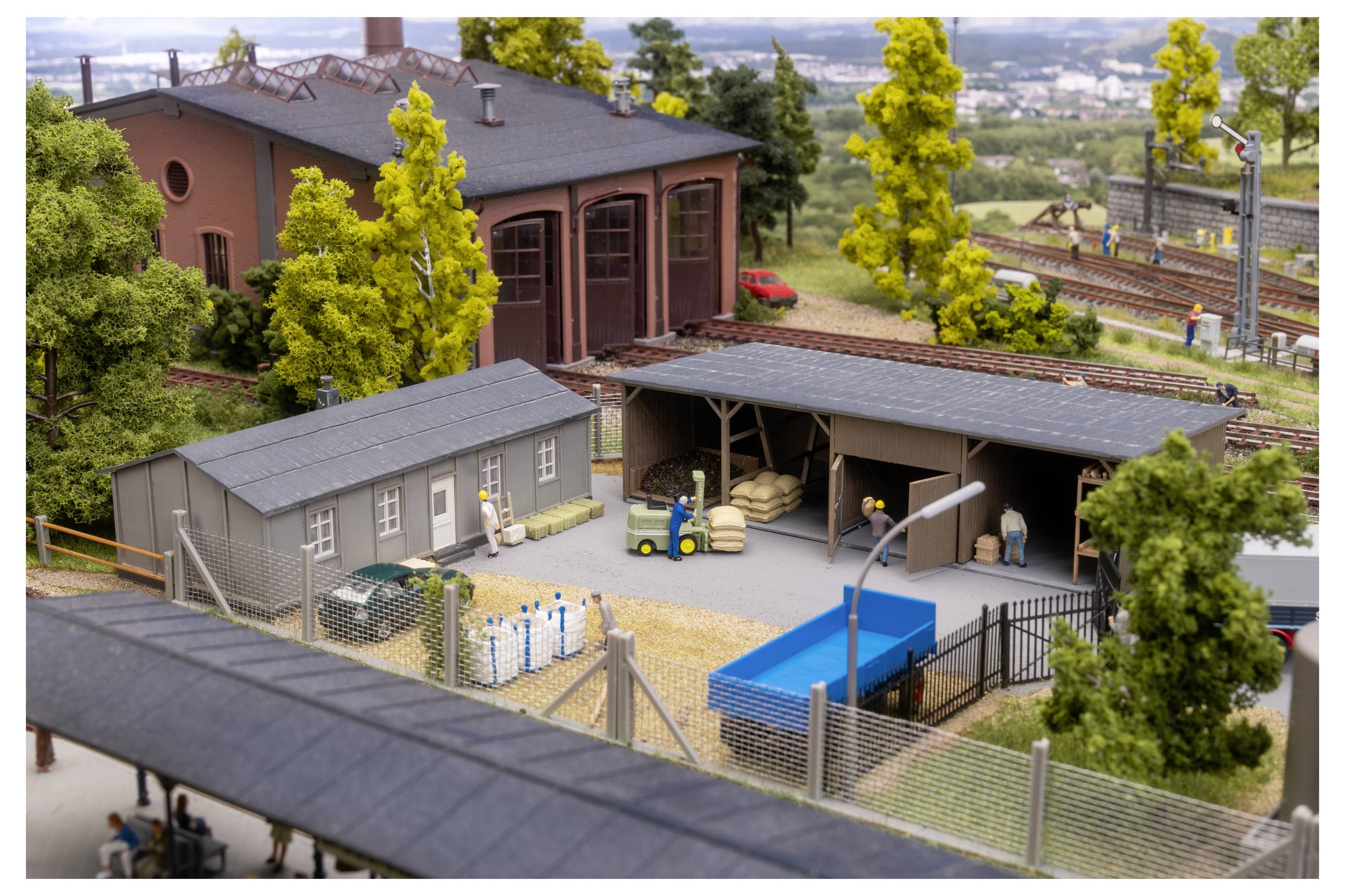 Modelleisenbahnanlage mit kleinen Industriegebäuden, eingezäuntem Gelände, Gleisen, Bäumen und winzigen Figuren in einer detaillierten Miniaturlandschaft.