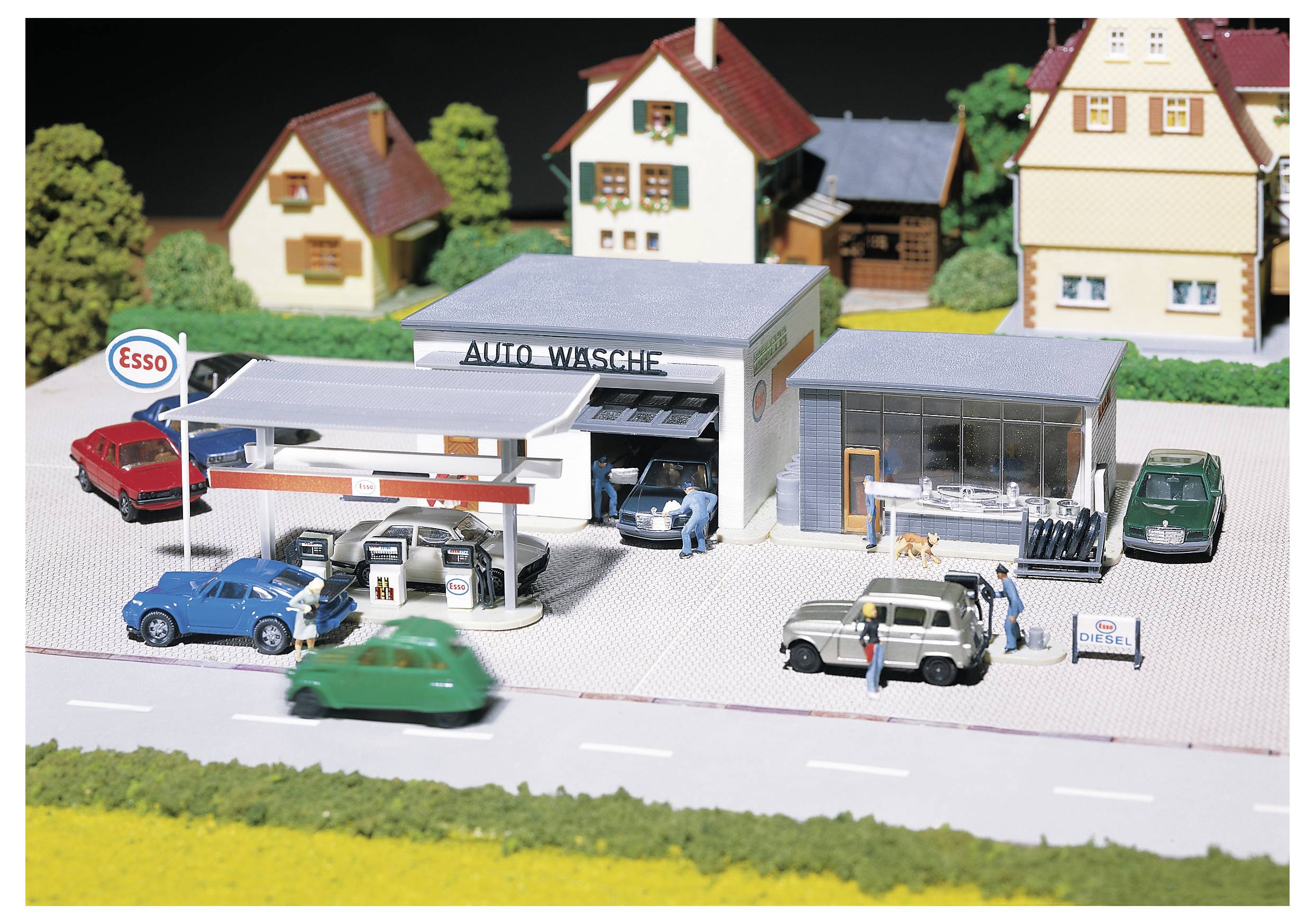Miniaturmodell einer Tankstelle und Autowäsche mit Autos, Zapfsäulen und kleinen Figuren in einer Dorflandschaft.