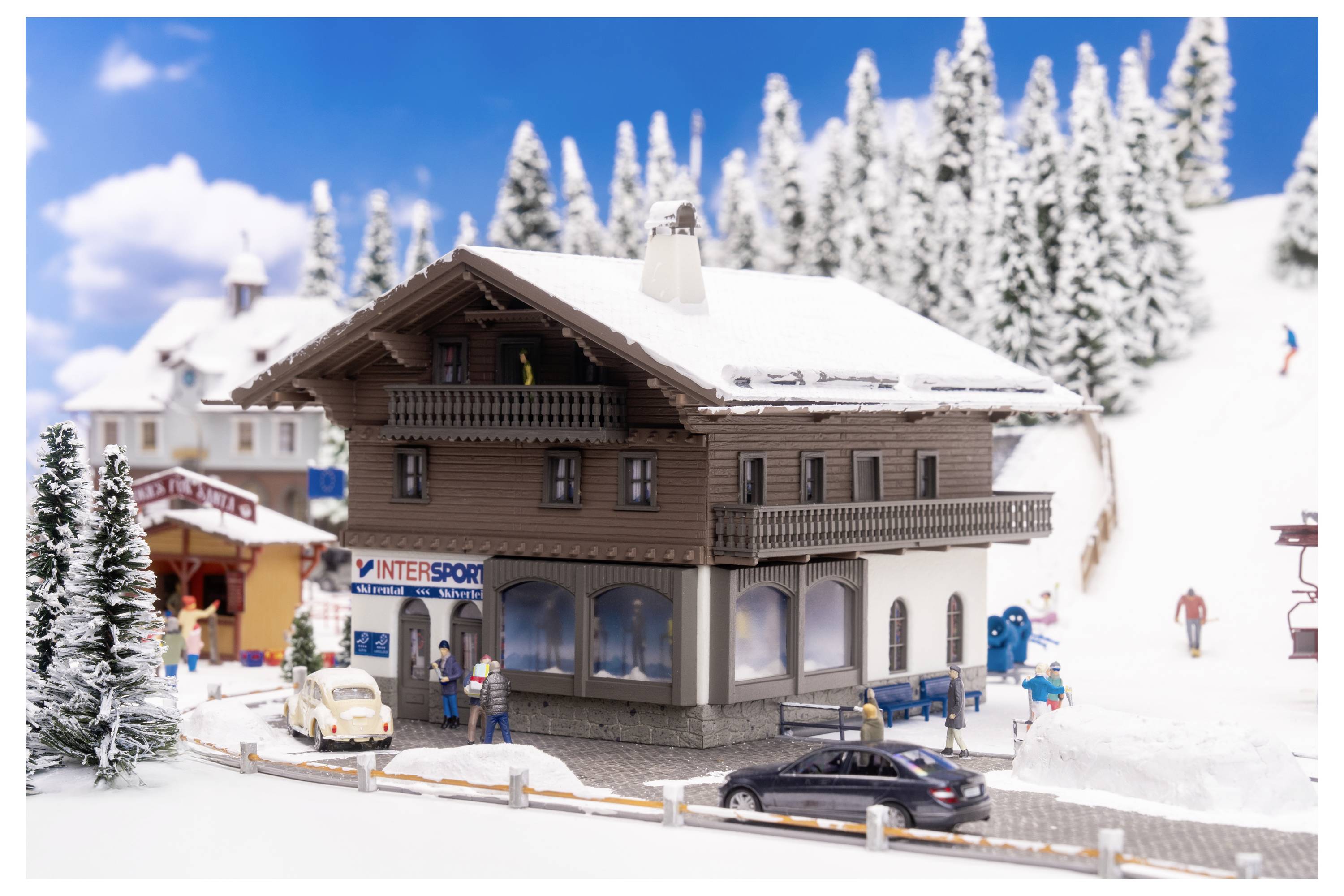 Ein winziges verschneites Skiort-Dorf mit Chaletgebäuden, geparkten Autos und Skifahrern an einem hellen Berghang im Winter.