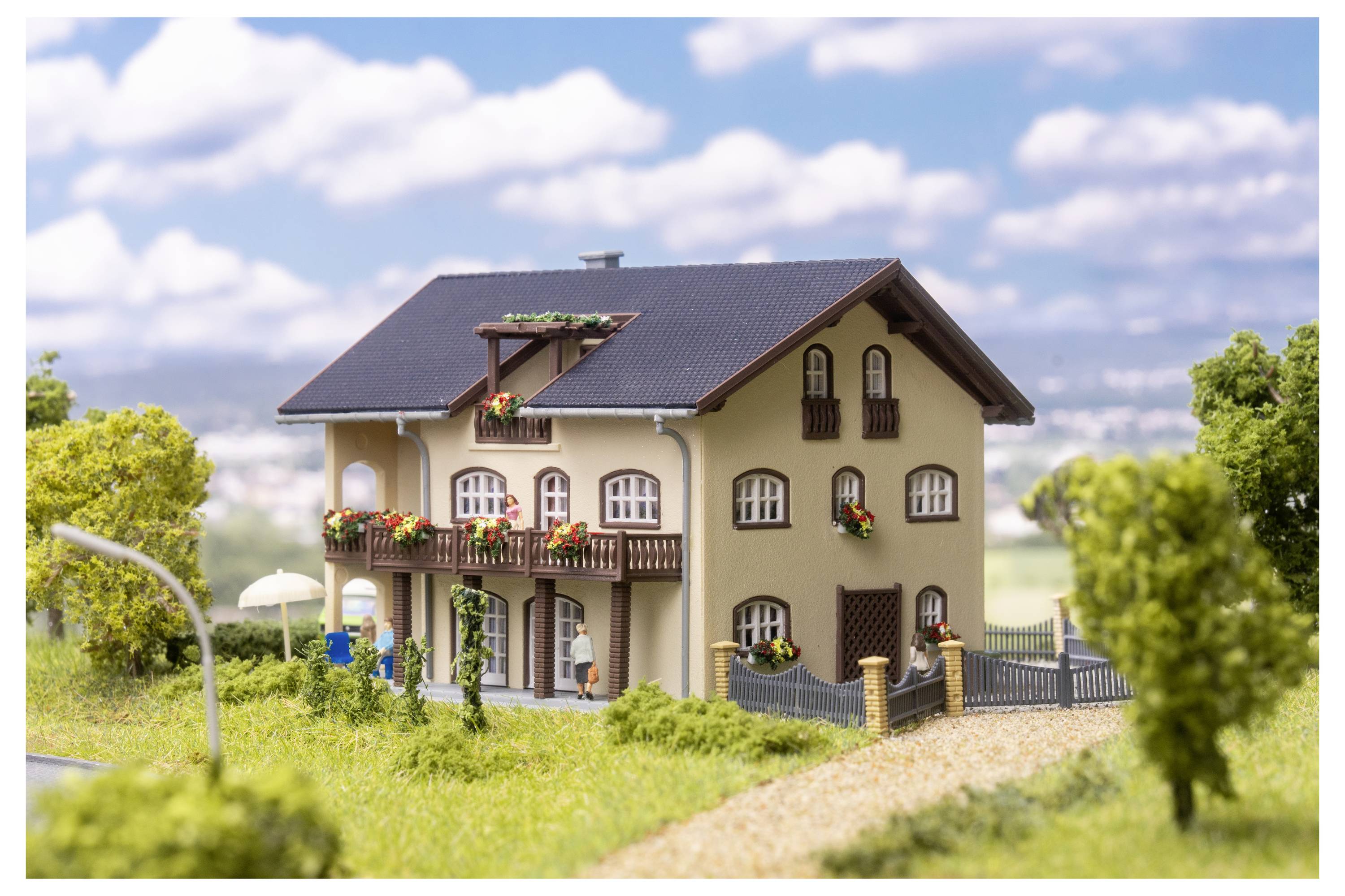 Miniaturmodell eines zweistöckigen Hauses mit Balkon, Garten und eingezäuntem Hof in einer ländlichen Landschaft.