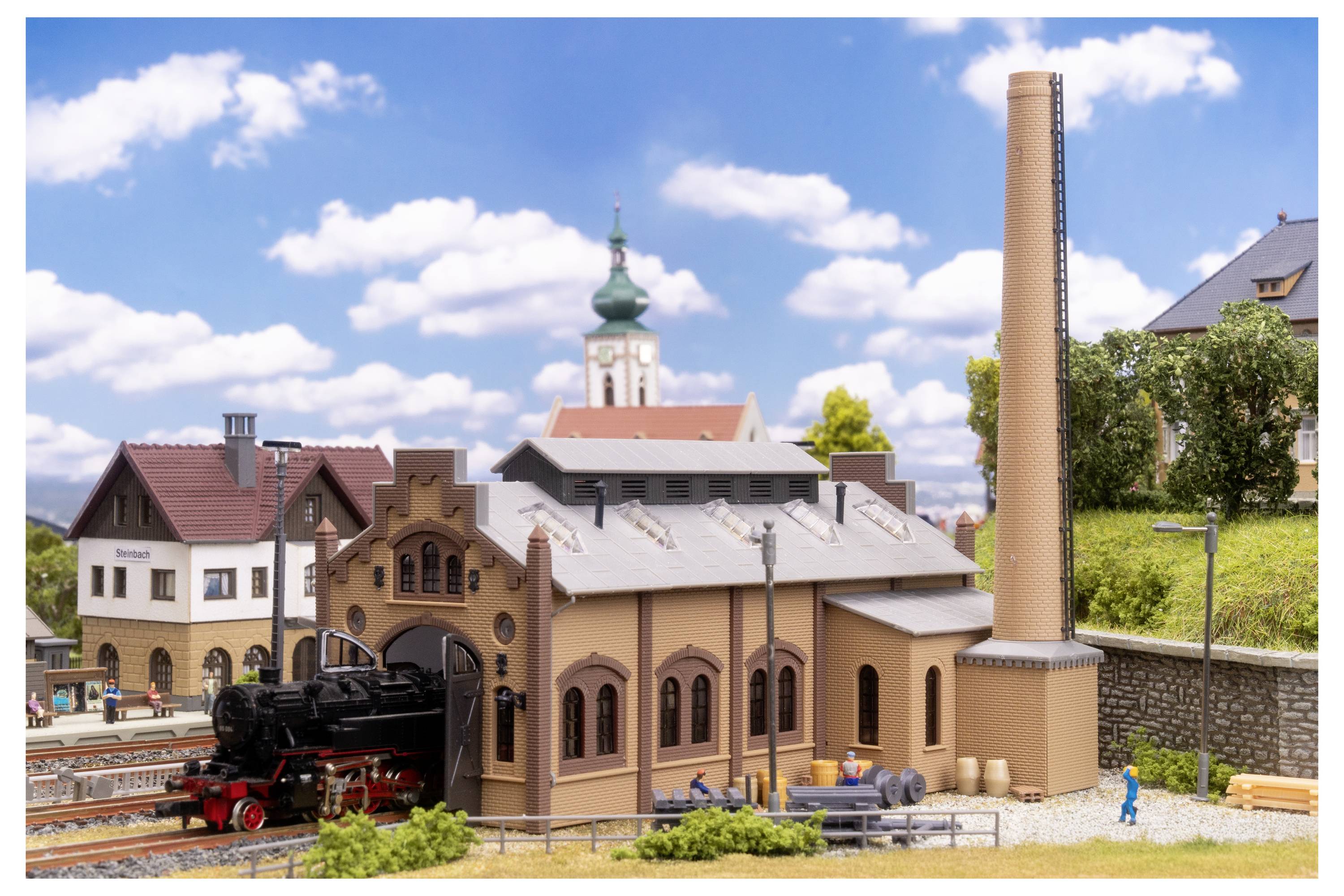 Modelleisenbahn-Szenerie mit einer Dampflokomotive neben einem gewölbten Depot, hohem Schornstein und Dorfgebäuden unter blauem Himmel.