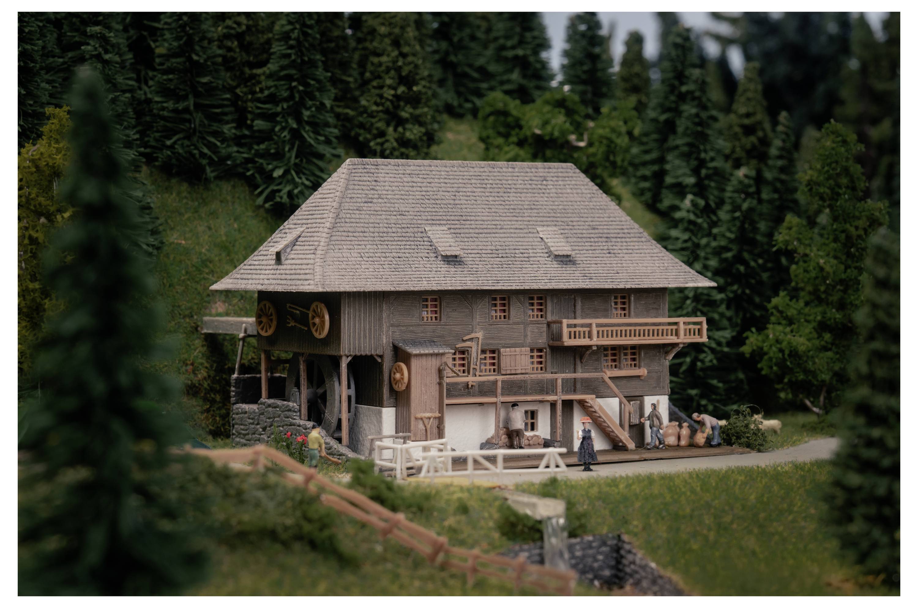 Ein Miniaturmodell eines rustikalen Holzhauses mit Balkonen und einem geneigten Dach, umgeben von dichtem Nadelwald und einer grünen Landschaft.