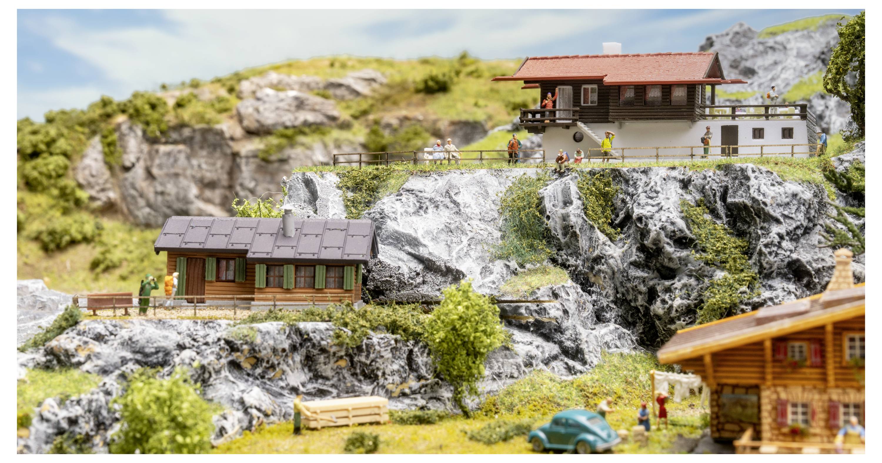 Miniatur-Bergdorfmodell mit Holzhütten auf felsigen Hängen, einem Kliffhaus, winzigen Figuren, Bäumen und einem kleinen blauen Auto.