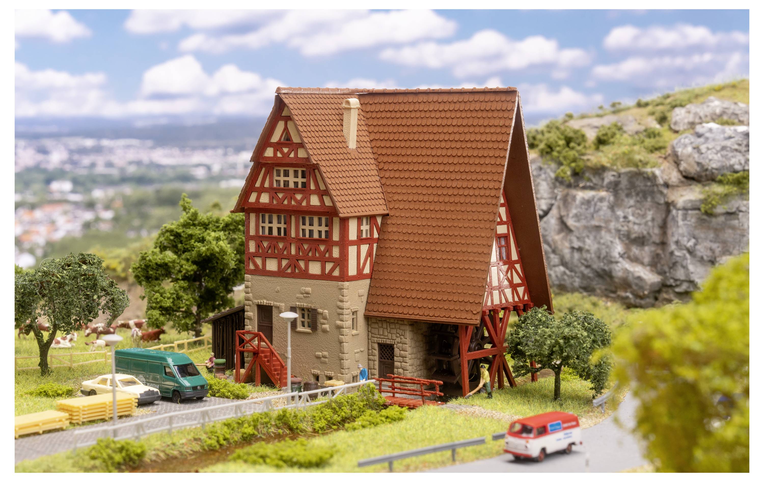 Detailliertes Modell eines Fachwerkhauses mit rotem Dach in einer ländlichen Miniaturlandschaft, mit Bäumen, Straße und kleinen Fahrzeugen in der Nähe.