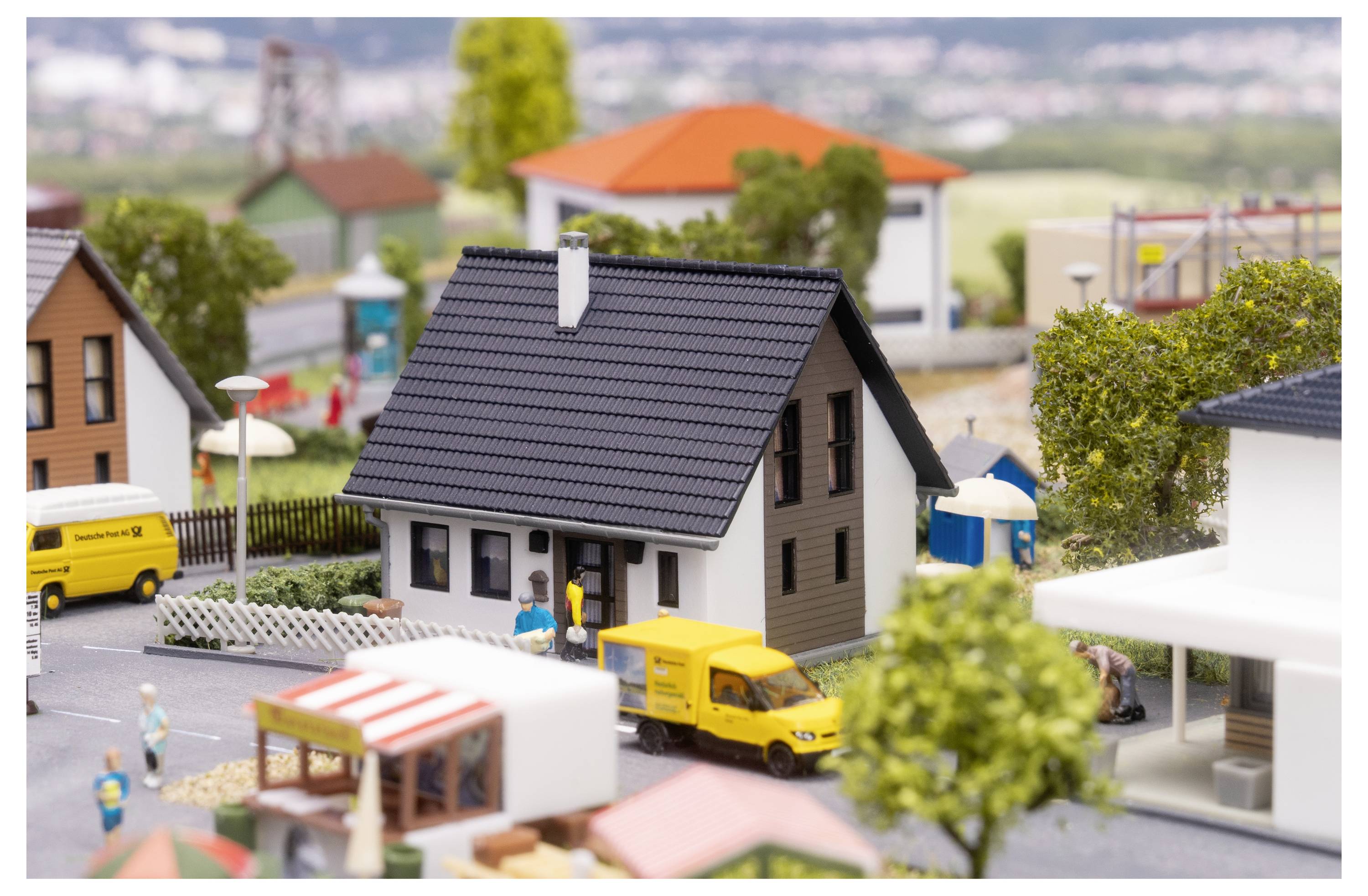 Miniaturmodell einer Wohnvorstadt mit einem zentralen grauen Haus, gelbem Lieferwagen und kleinen Figuren auf einer Straße.
