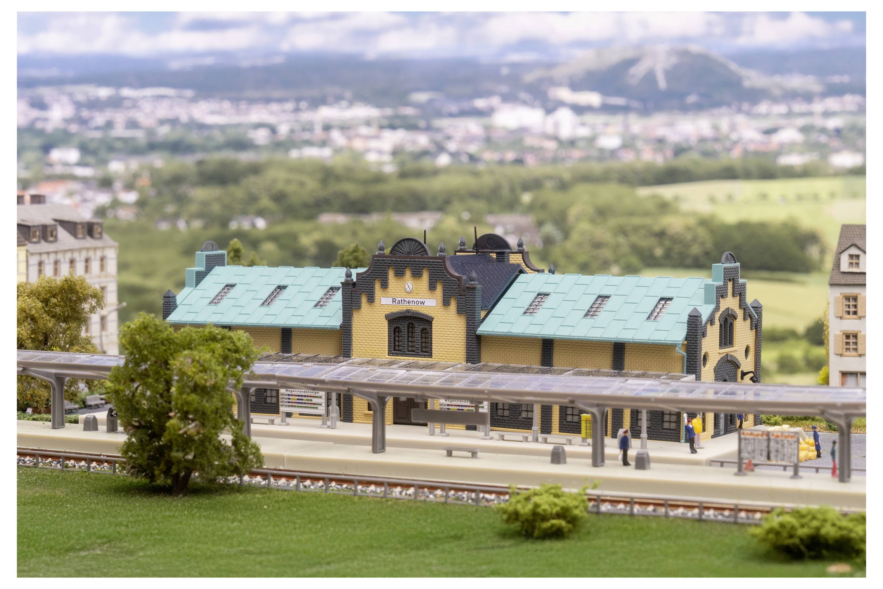 Miniaturmodell eines Bahnhofs mit türkisfarbenem Dach und Bahnsteig, eingebettet in ein gestaltetes Modelldorf mit entfernten Hügeln im Hintergrund.
