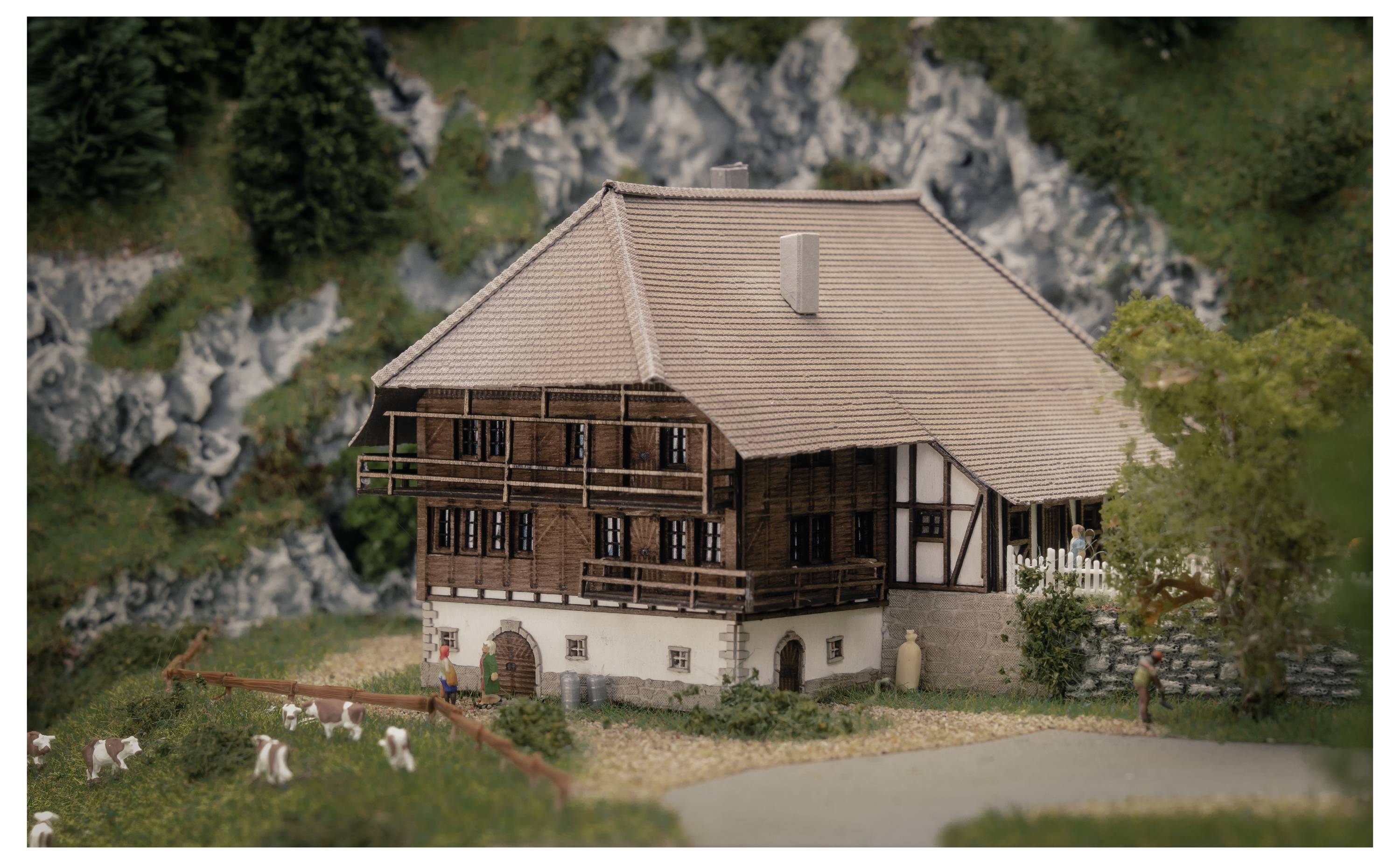 Ein Modell eines rustikalen zweistöckigen Bauernhauses mit Ziegeldach, das neben einem eingezäunten Rasenplatz mit kleinen Weidetieren und einer felsigen Hügellandschaft im Hintergrund positioniert ist.