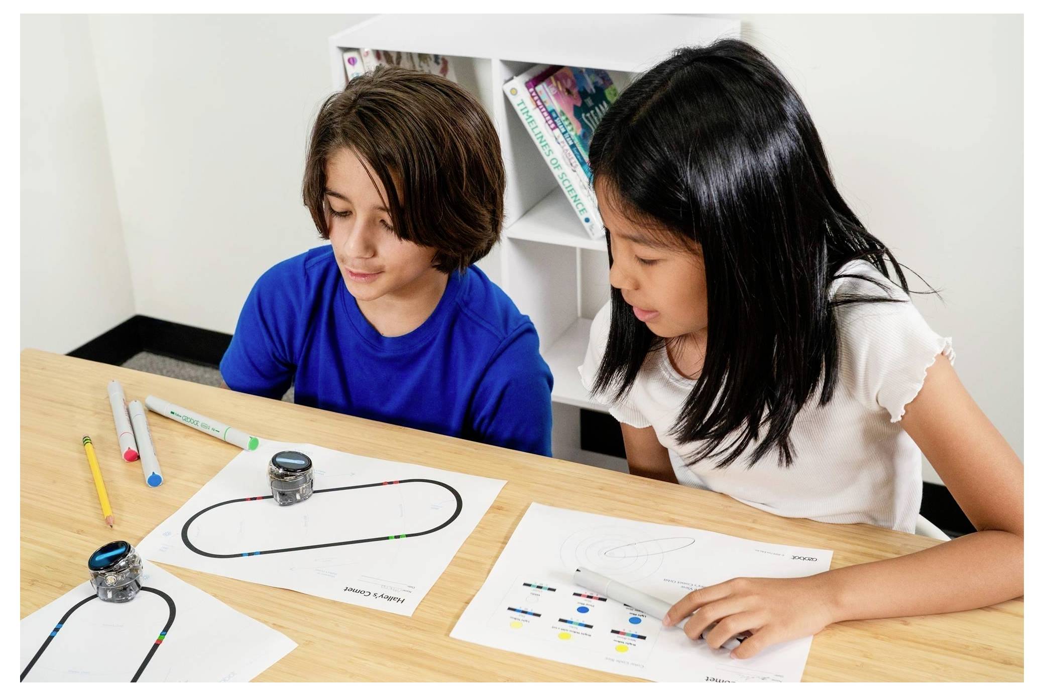 Zwei Kinder studieren ein Papierlabyrinth oder Spurenpuzzle an einem Tisch und verwenden dabei kleine Roboter oder Marker während einer Klassenraumaktivität.