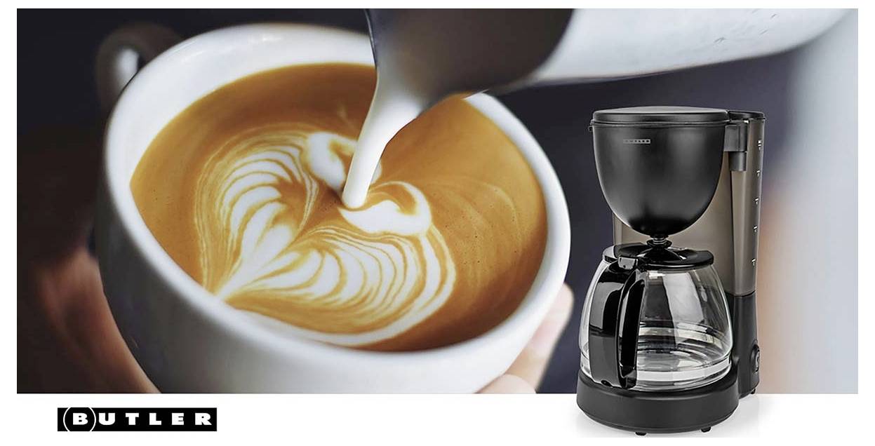 Kaffeemaschine mit glassigem Krug neben einem Latte, in den Milch gegossen wird, wodurch Latte Art in einer weißen Tasse entsteht; Butler-Marke abgebildet.