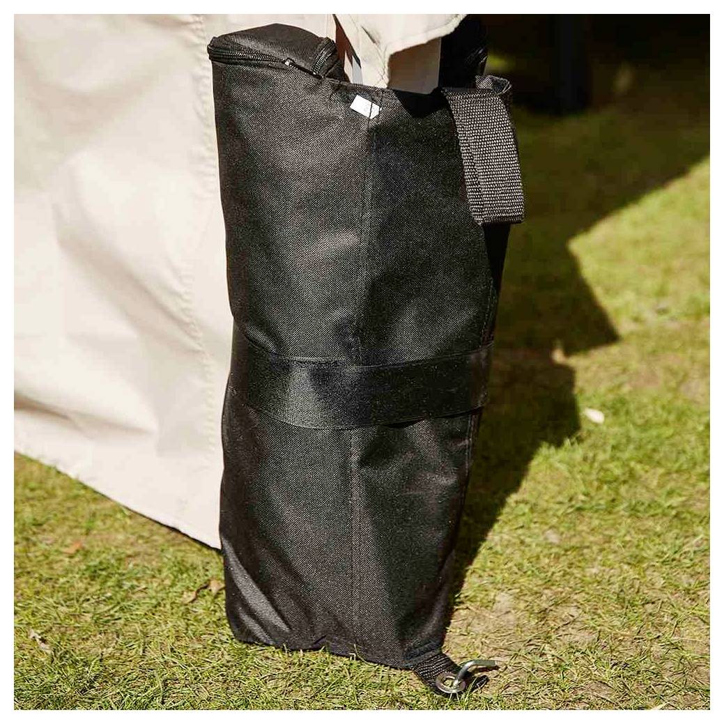 Schwarze zylindrische Outdoor-Tasche mit Riemen und Kordelzug sitzt auf Gras neben einer hellfarbigen Zeltmauer.