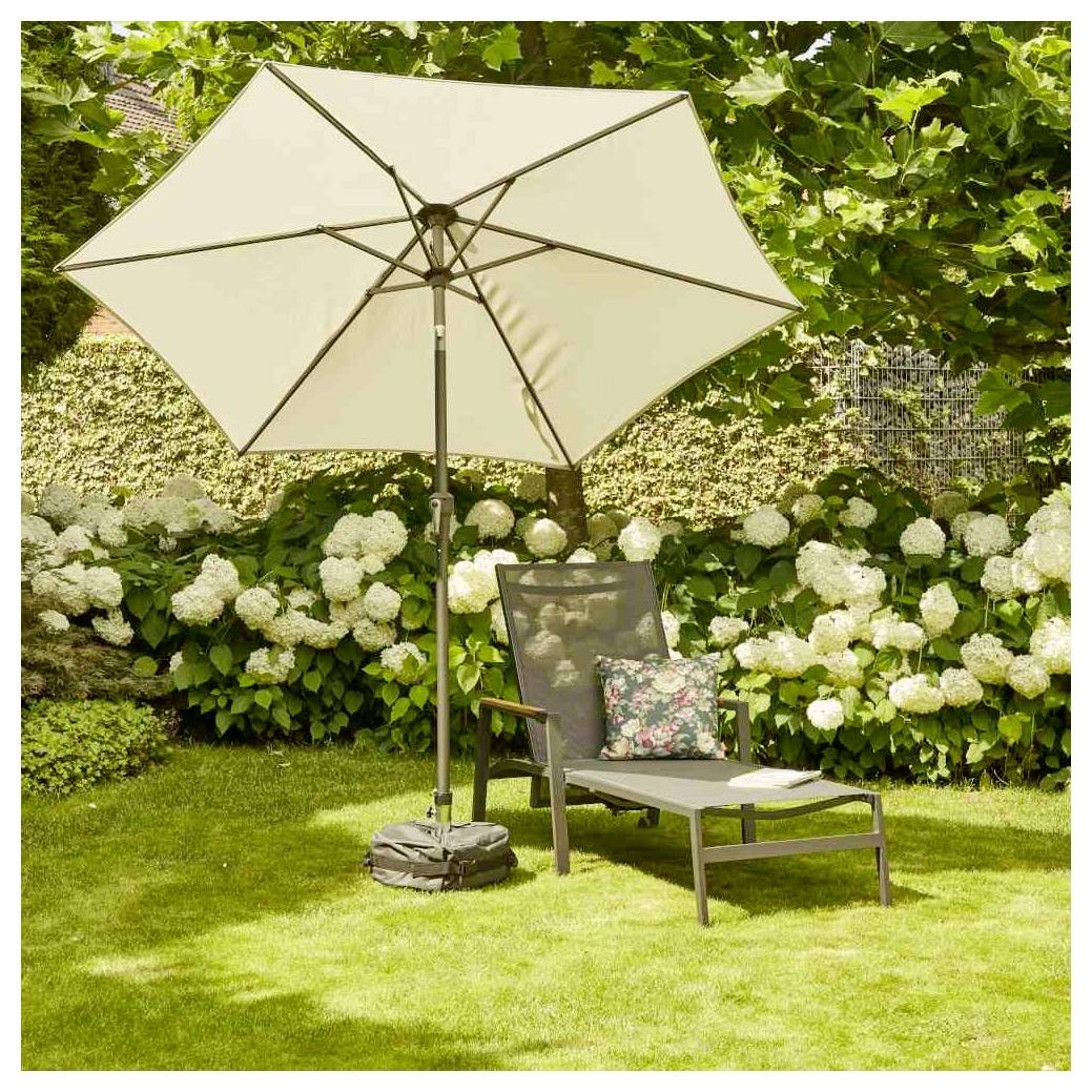 Loungesessel für die Terrasse mit geblümtem Kissen unter einem großen beigefarbenen Gartenschirm auf einem Rasen, mit weißen Hortensien und Grünflächen im Hintergrund.