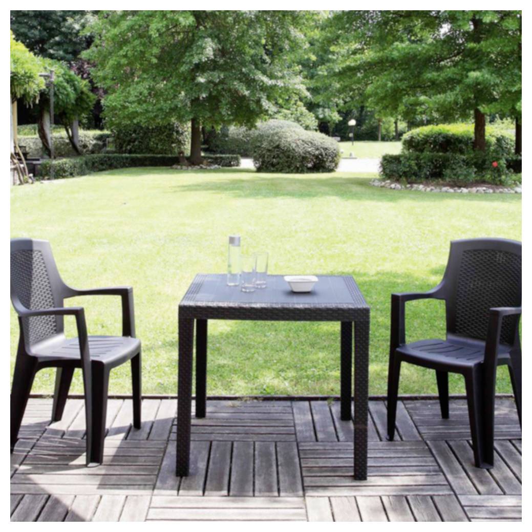 Zwei leere Stühle flankieren einen kleinen Tisch auf einer hölzernen Terrasse, die auf einen grasbewachsenen Garten blickt.