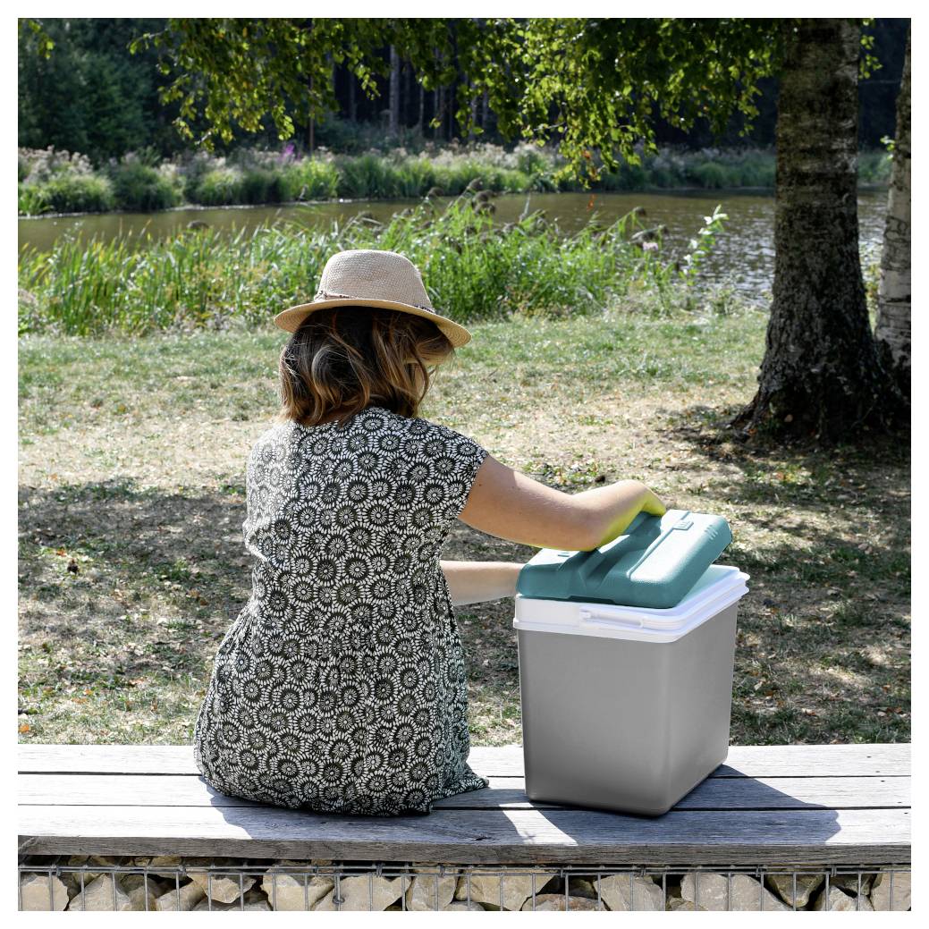 Eine Frau mit Hut sitzt auf einer Bank neben einem Teich und öffnet eine Kühlbox oder Lagerkiste im Freien in einem Park.