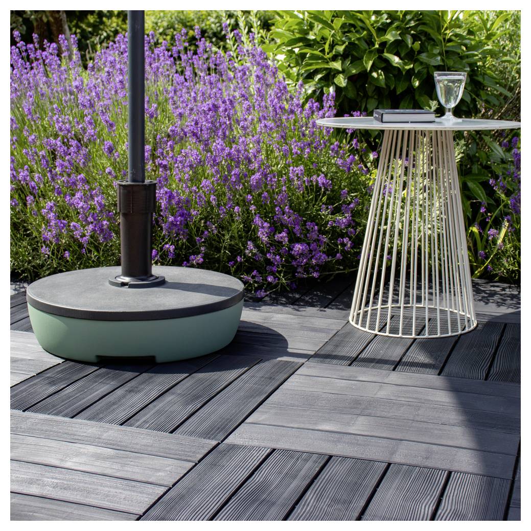 Moderne Terrasse mit einem runden Außentisch aus Glas und einer Heizerbasis neben violetten Blüten und grünen Sträuchern auf einem Holzbelag.