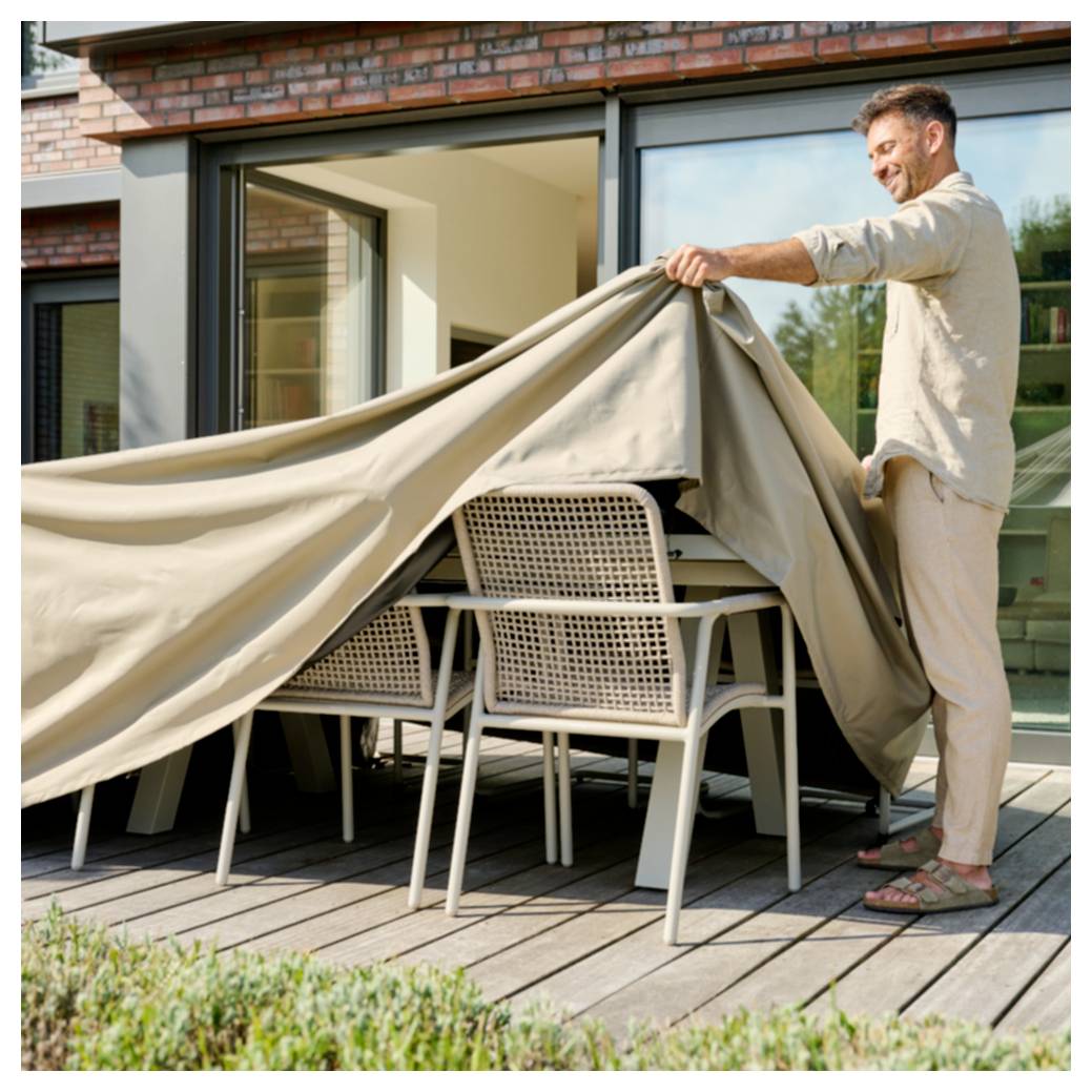Ein Mann auf einer Terrasse, der eine große Abdeckung von Außenmöbeln zum Speisen auf einem Holzdeck außerhalb eines Hauses abzieht.
