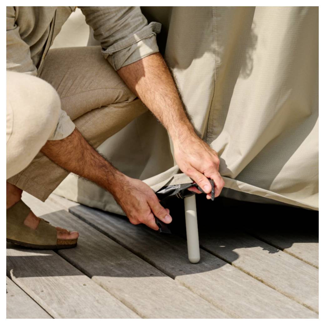 Eine Person hockt sich hin, um eine beige Outdoor-Möbelschutzhülle um ein Stuhlbein auf einem Holzdeck zu befestigen oder zu sichern.