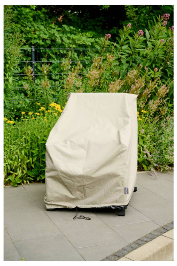 Eine beige Schutzabdeckung liegt über einem Rollstuhl im Freien oder einem Grill auf einer Terrasse, mit Gartenpflanzen im Hintergrund.