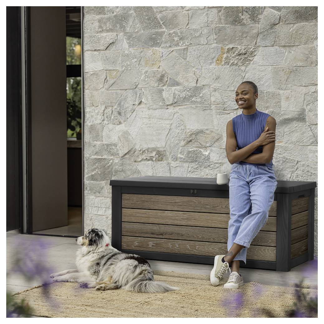Lächelnde Frau in ärmellosen blauem Top und Jeans lehnt sich gegen einen Terrassenschrank neben einem Hund, der auf einem Teppich vor einer Steinmauer liegt.