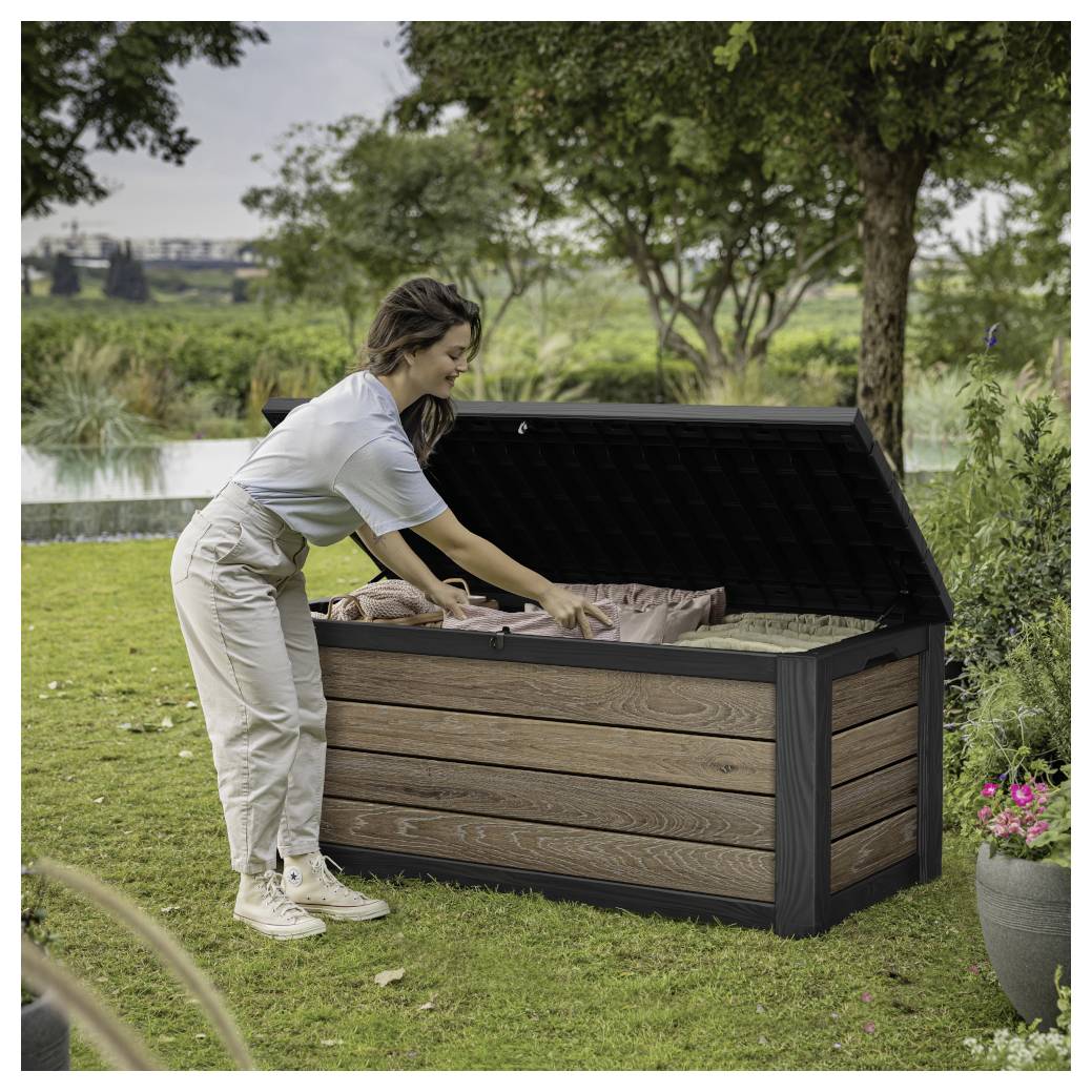 Eine Frau packt Gegenstände in eine große Außenlagerungsbox in einem grasbewachsenen Garten in der Nähe eines Teiches.
