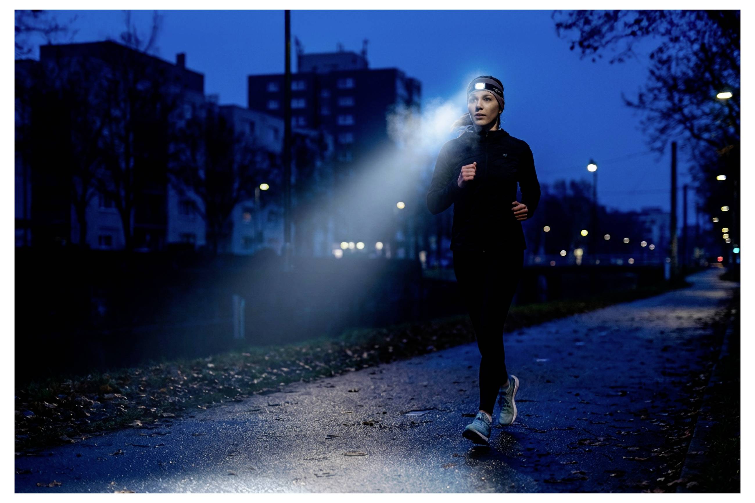 Frau beim Joggen nachts auf einem nassen Stadtpfad, die eine Stirnlampe trägt, die einen hellen Strahl nach vorne wirft.