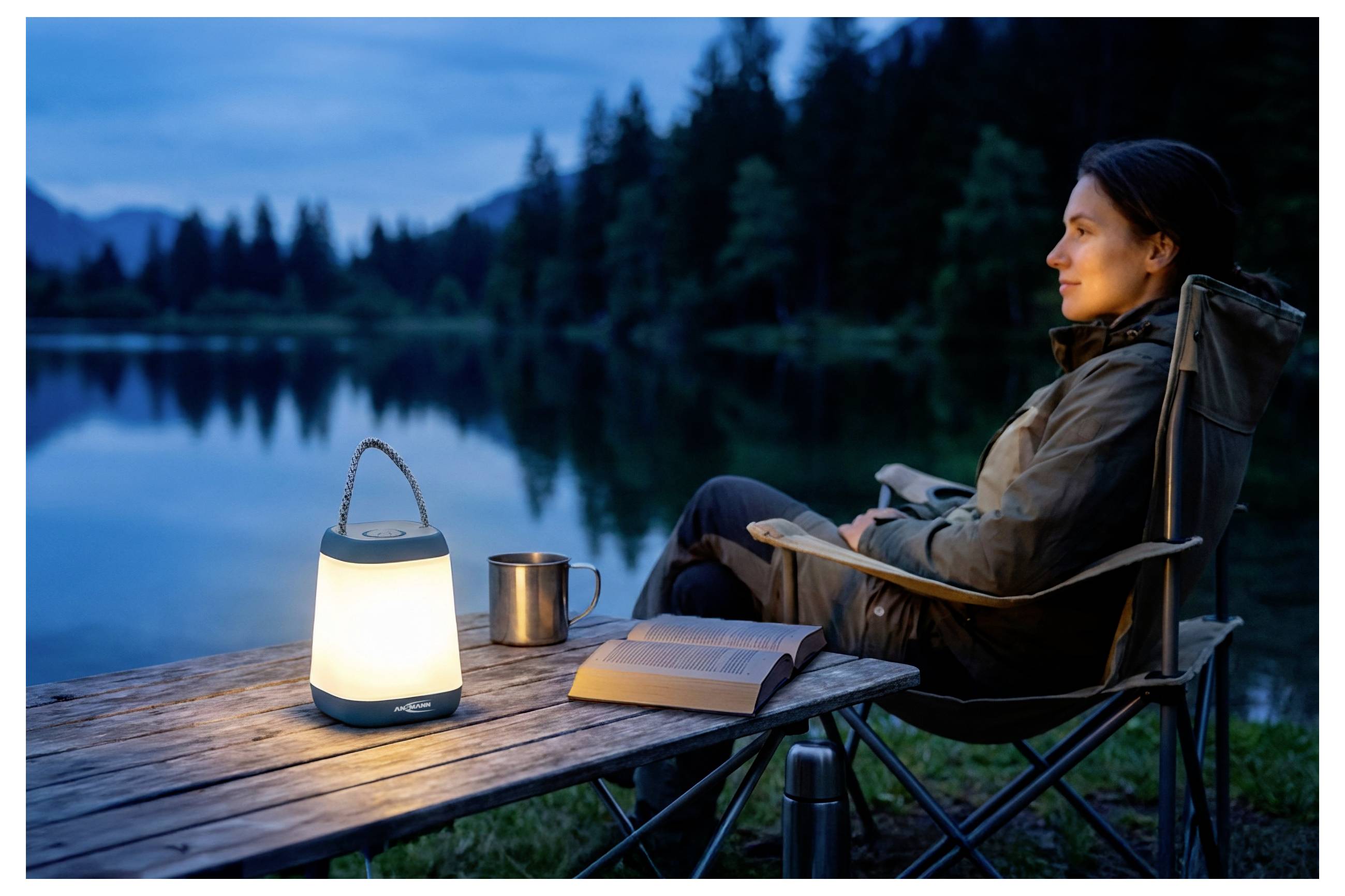 Frau, die sich in einem Campingstuhl neben einem See in der Dämmerung entspannt, mit einer brennenden Laterne, einer Tasse und einem aufgeschlagenen Buch auf einem Holztisch.
