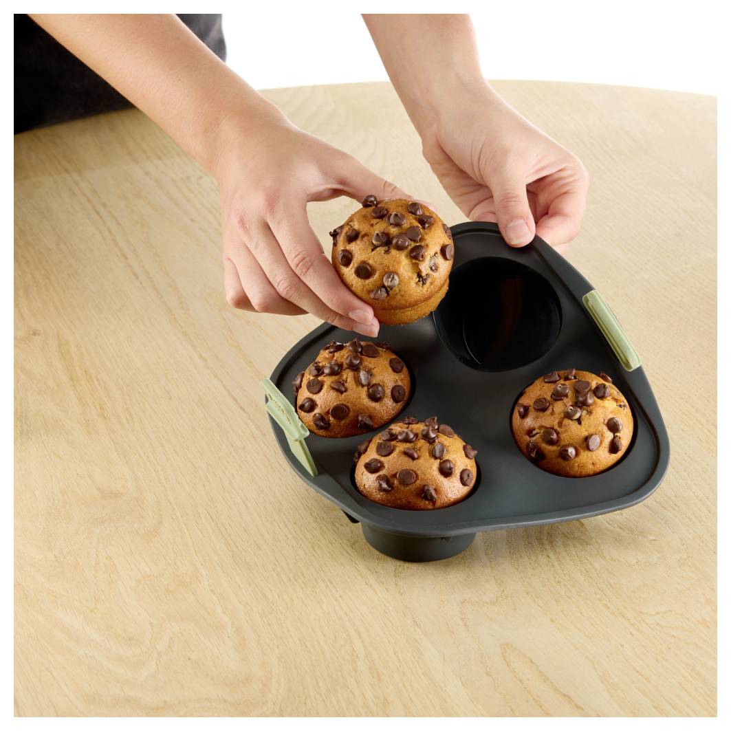 Hände legen einen Schokoladenstückchen-Muffin in eine schwarze Backform für vier Stück auf einem Holztisch.