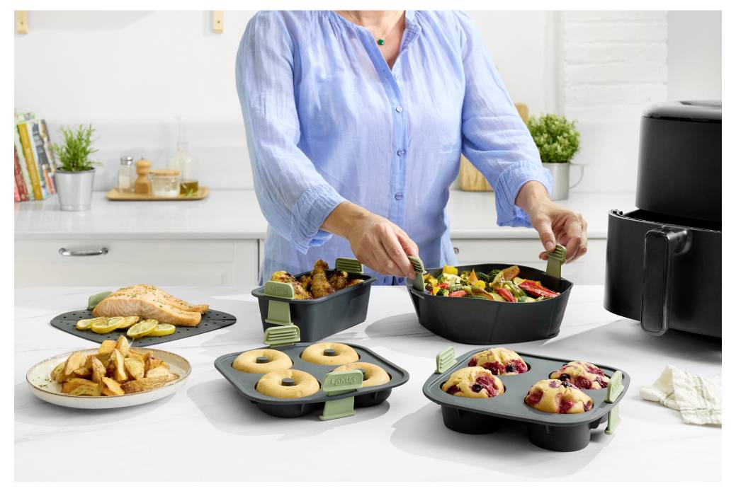 Person in einem blauen Hemd serviert Essen aus schwarzen Meal-Prep-Behältern mit geröstetem Huhn, Kartoffeln, Salat, Donuts und Beerengebäck auf einer Küchentheke neben einer Heißluftfritteuse.