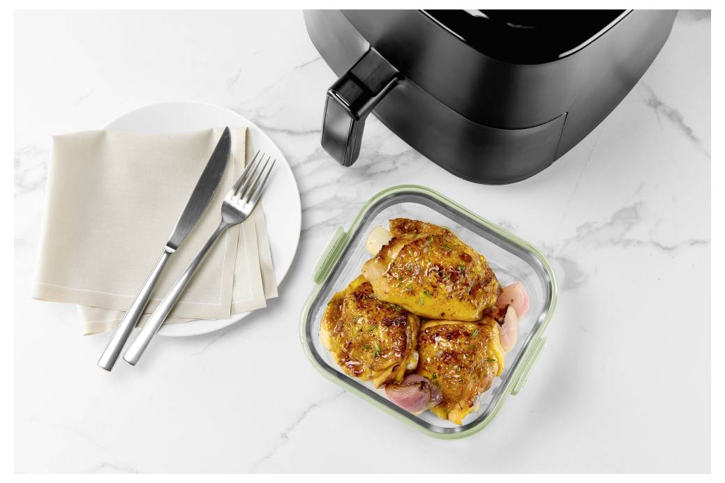 Meal-Prep-Behälter mit gekochtem Hähnchen in einer Heißluftfritteuse, daneben ein Teller mit Serviette und Besteck auf einer Marmortheke.