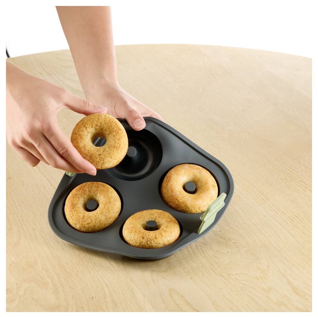 Hände legen einen gebackenen Donut in eine schwarze Donut-Form mit sechs Fächern auf einem Holztisch; vier Donuts befinden sich bereits im Tablett.