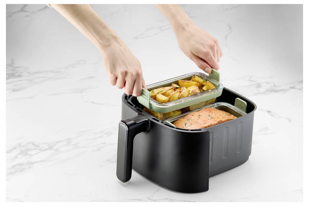Hände, die einen Glasmahlzeitbehälter mit gerösteten Kartoffeln in eine schwarze elektrische Lunchbox mit gekochtem Lachs auf einer weißen Arbeitsplatte stellen.