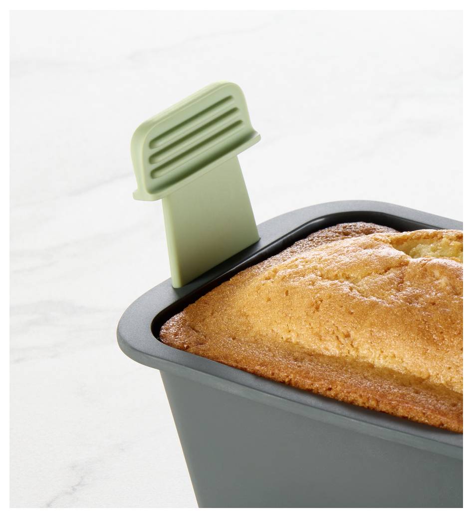 Frisch gebackener Kastenkuchen in einer dunkelgrauen Kastenform mit grünem Silikongriffband, gezeigt auf einer weißen Oberfläche.