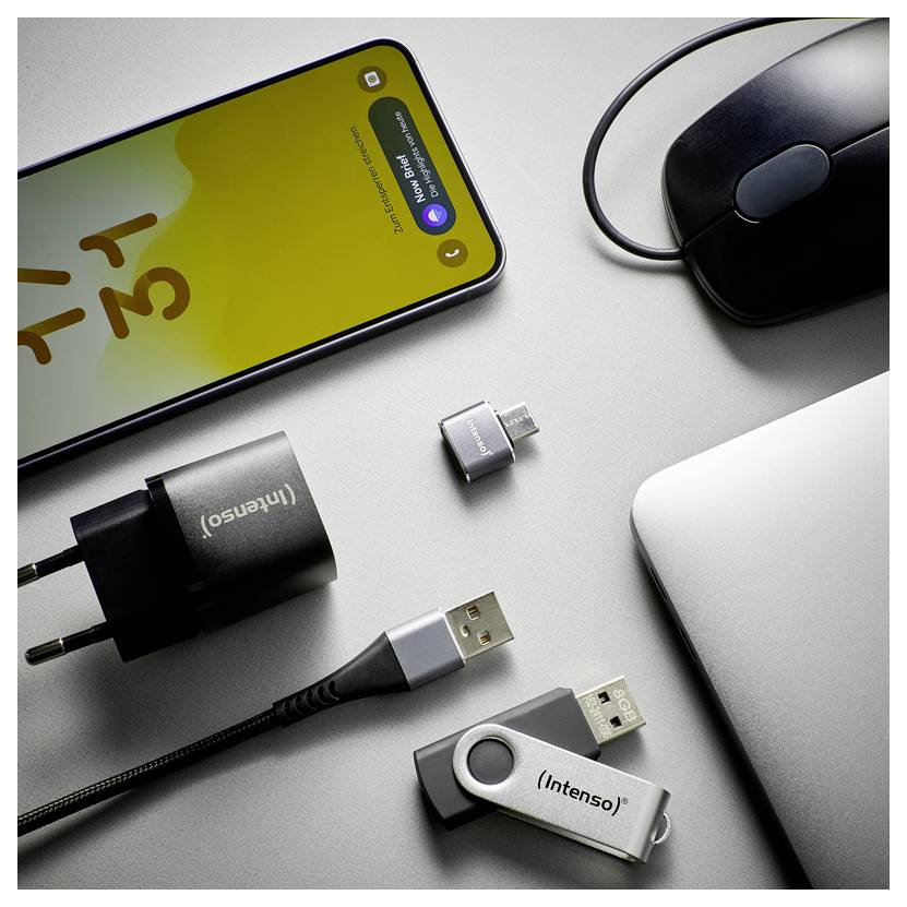 Ein Smartphone, eine Maus, eine Laptop-Ecke, ein Ladegerät und zwei USB-Adapter sind auf einem grauen Schreibtisch angeordnet, wobei ein Adapter in ein Kabel eingesteckt ist.