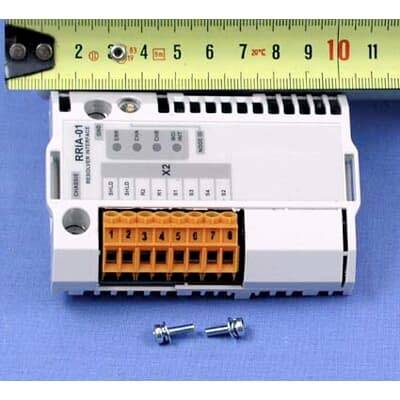Weißes elektronisches Modul auf einer blauen Oberfläche mit einem Messstab darüber und zwei kleinen Schrauben darunter, die seine Größe zeigen.