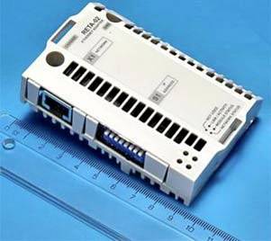 Weißes elektronisches Modul oder Sensor mit Lüftungsöffnungen und Anschlüssen, platziert auf einer blauen Messmatte mit Linealmeter.