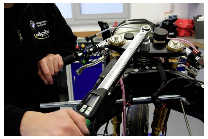 Mechaniker stellt das Vorderteil und die Lenker eines Motorrads in einer Werkstatt ein, mit sichtbarem BMW-Logo auf dem Hemd der Person.