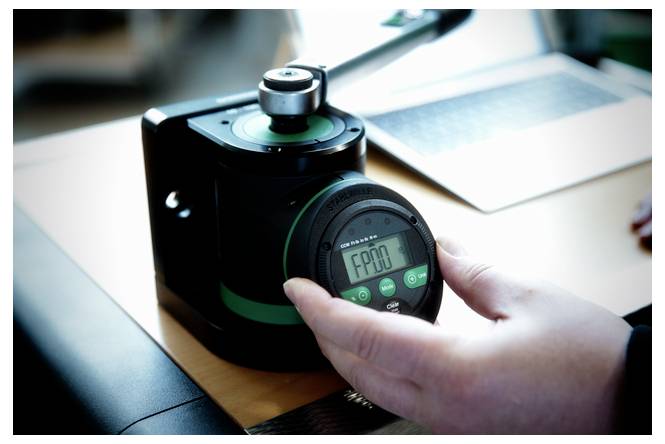 Hand, die eine Stoppuhr neben einer schwarzen Kaffeemühle auf einem Schreibtisch mit einem Laptop im Hintergrund hält.