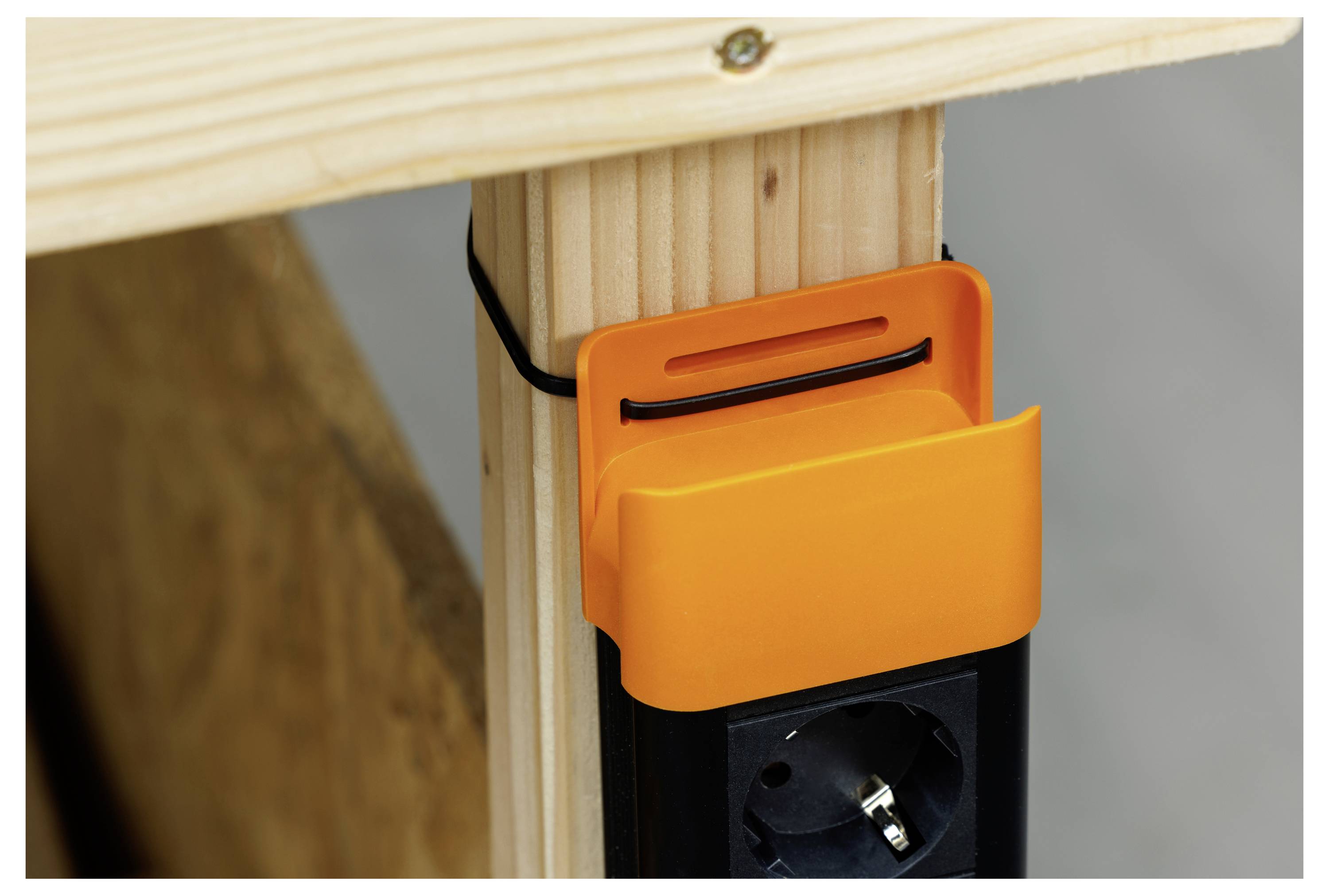 Orange Kabelorganizer-Clips befestigen ein schwarzes Stromkabel an einem Holzpfosten über einer Steckdose.