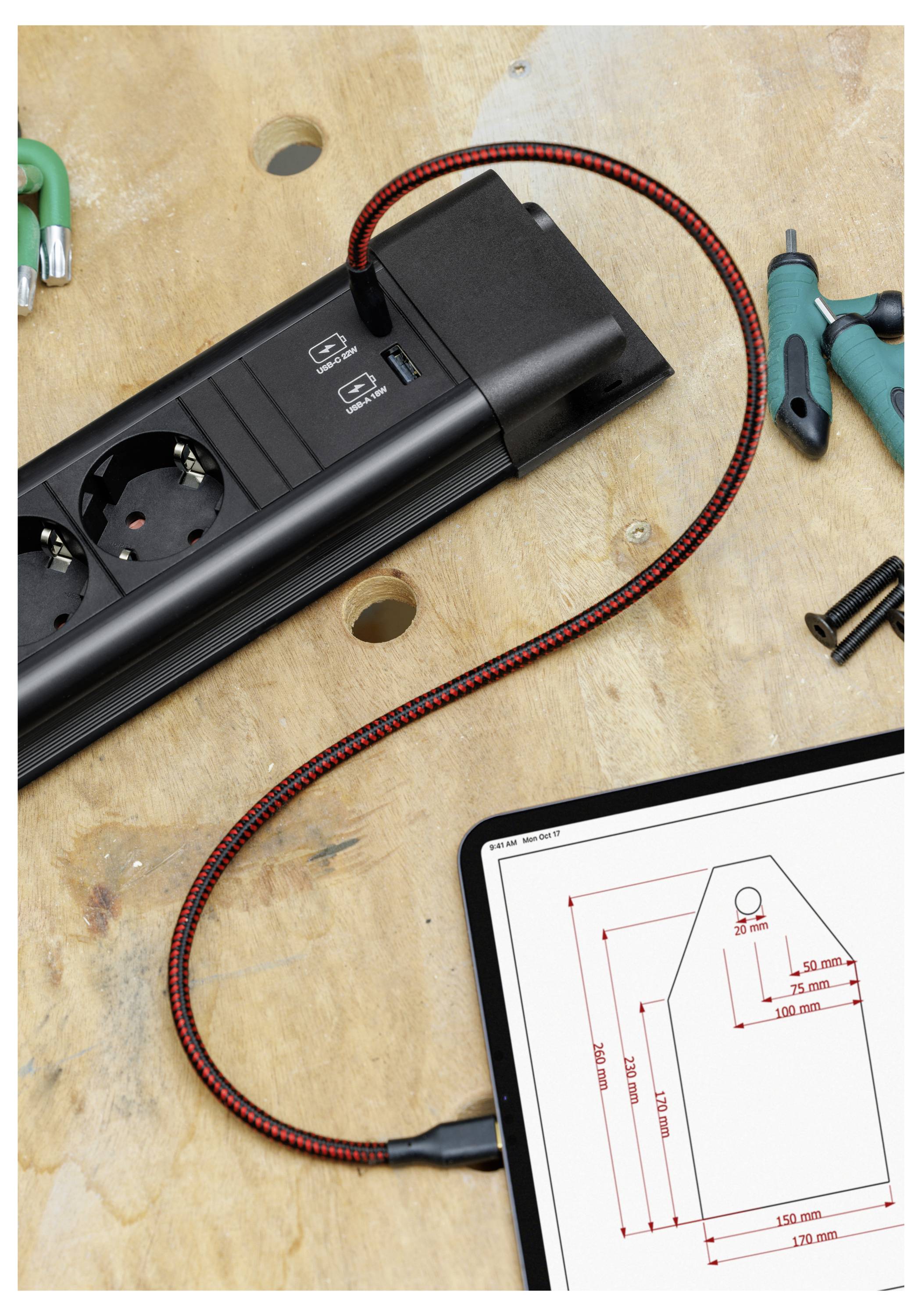 Ein schwarzes Steckerleistengerät mit rot-schwarzem Kabel liegt auf einer Werkbank neben Werkzeugen und einem Tablet, das eine technische Maßzeichnung zeigt.