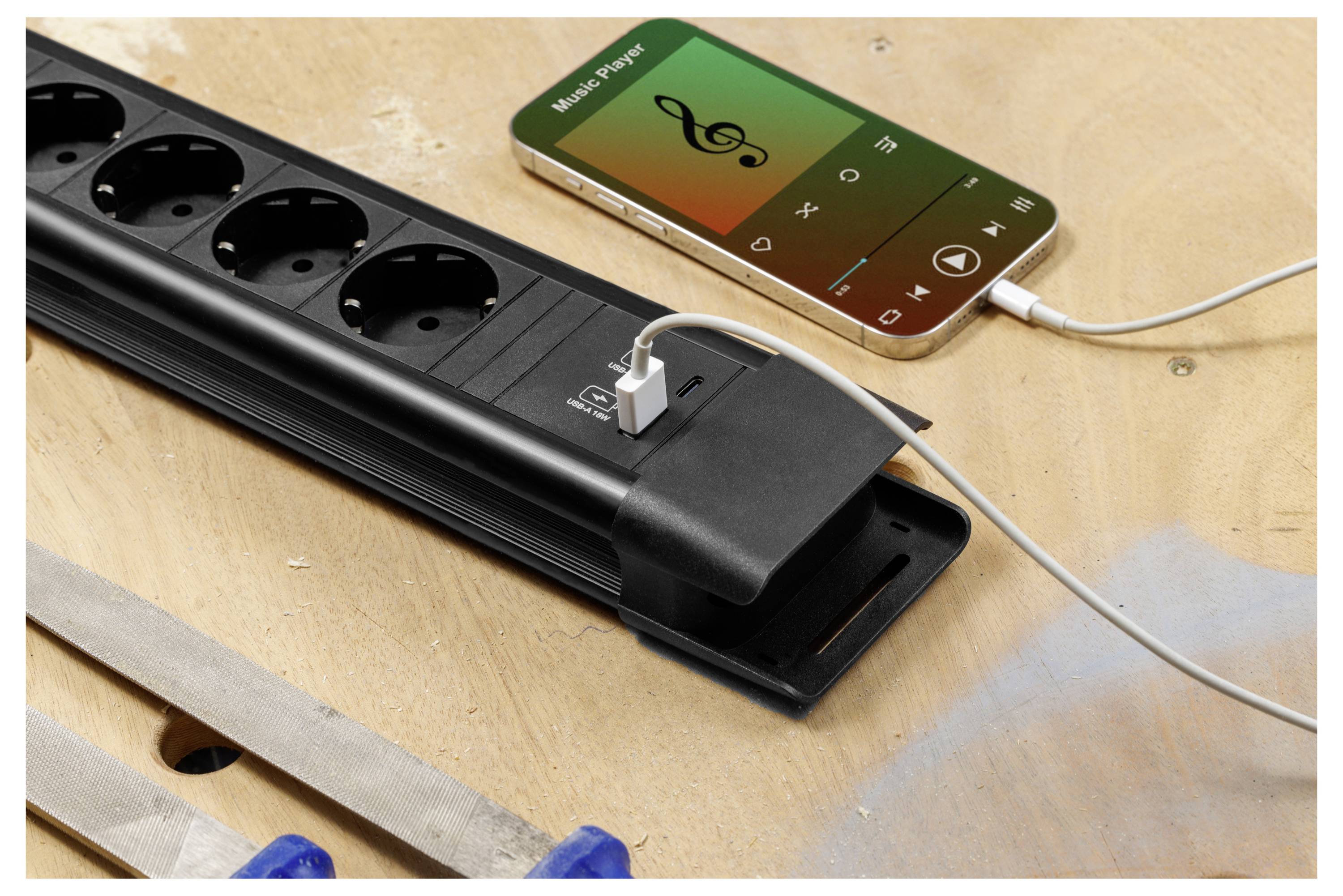 Mobiltelefon, das Musik abspielt, während es über ein Kabel mit einer Steckerleiste mit USB-Ladegerät auf einer Werkbank verbunden ist und Laden und Musikwiedergabe anzeigt.