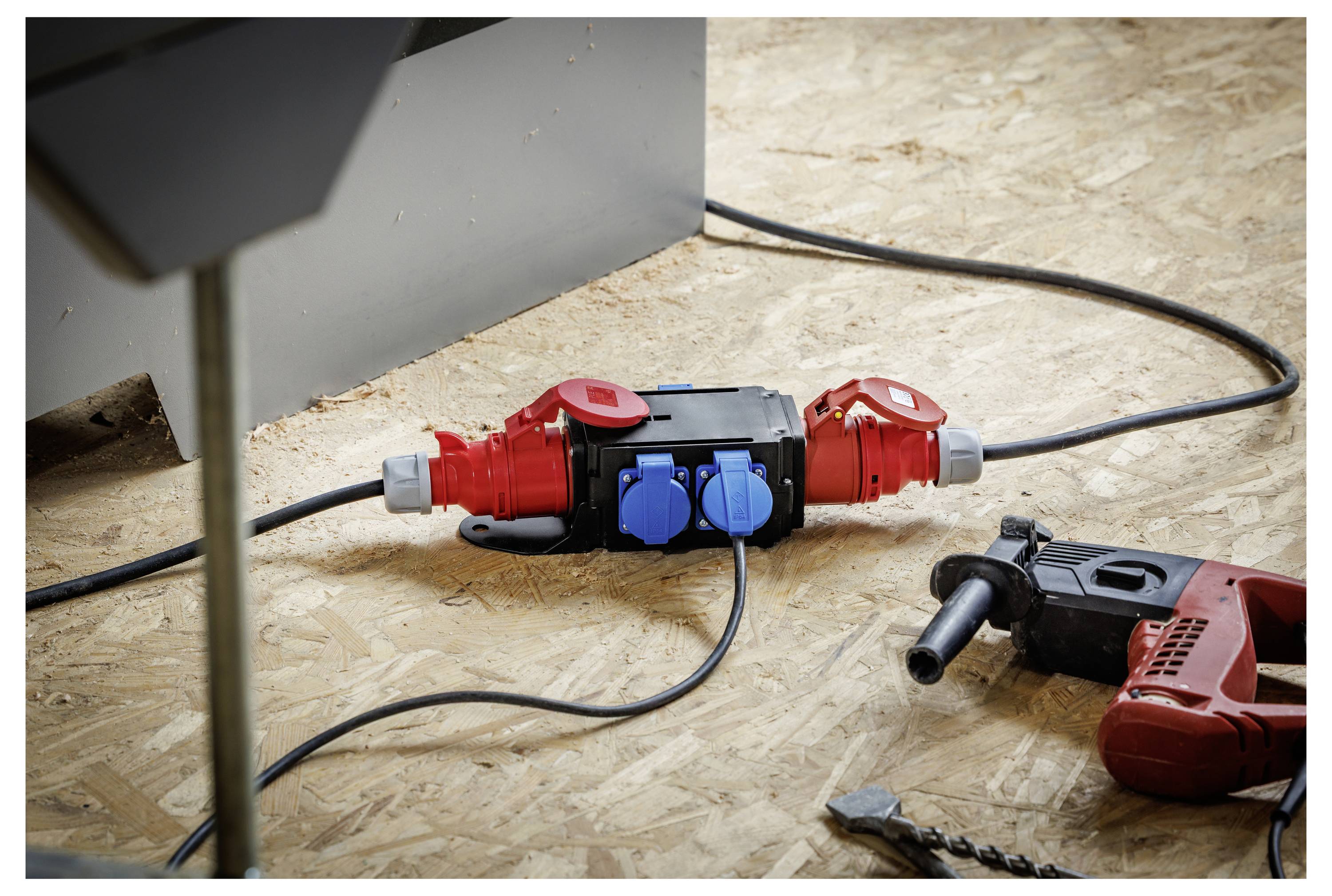 Tragbarer elektrischer Stromverteiler mit zwei roten Steckdosen und blauen Buchsen auf einer Sperrholzarbeitsfläche, mit Kabeln und einer Bohrmaschine in der Nähe in einer Werkstattumgebung.