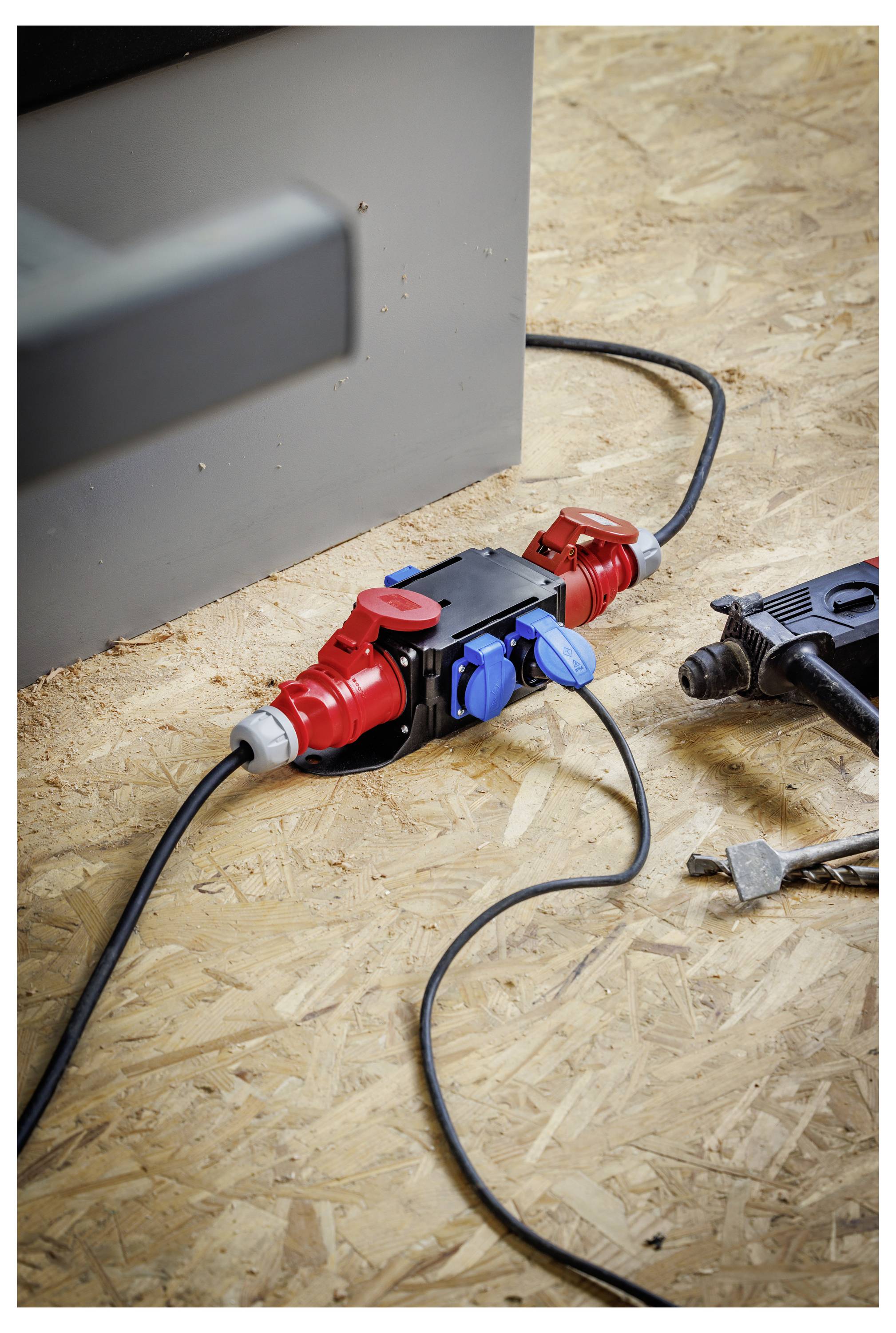 Ein roter und blauer Outdoor-Stromverteiler auf einem Sperrholzboden mit schwarzen Kabeln, die neben einer Wand eingesteckt sind.