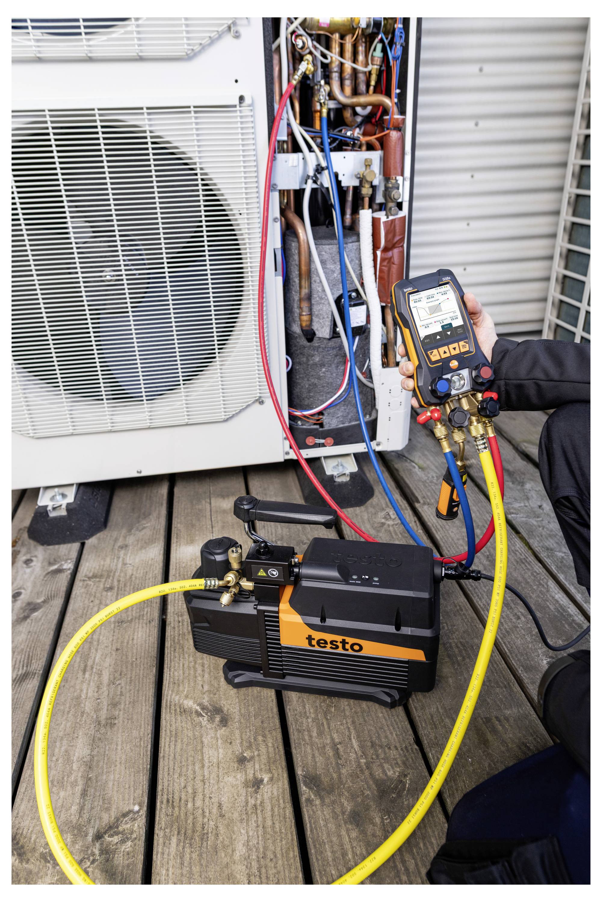 Techniker testet eine Außenklimaanlage mit einem Testo-Manometer und Schläuchen, die an den freigelegten Kältemittelleitungen der Anlage angeschlossen sind.