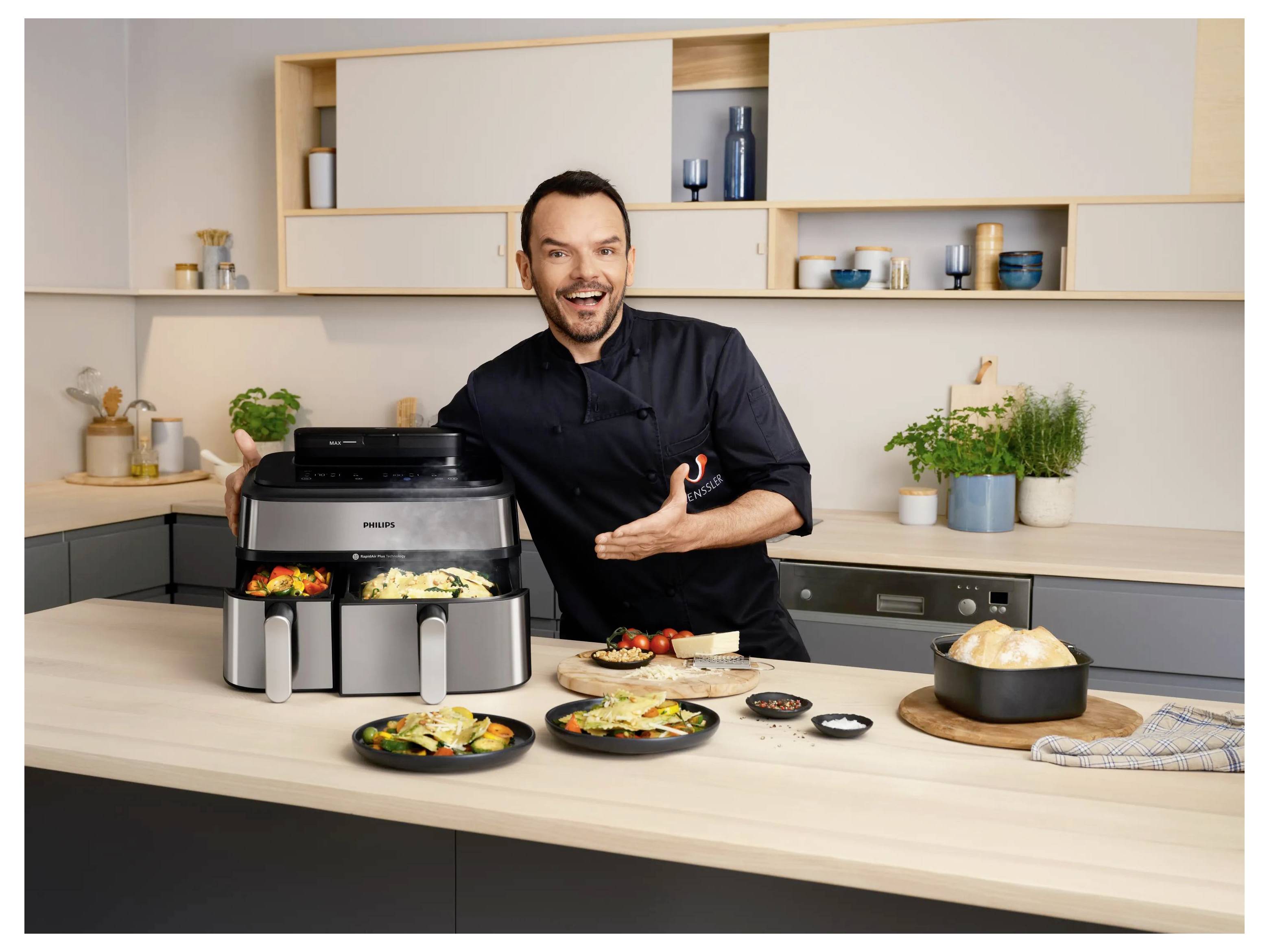 Ein lächelnder Mann präsentiert eine Philips-Fritteuse mit gekochtem Essen in einer modernen Küche, umgeben von angerichteten Tellern und einer Schüssel mit Reis.
